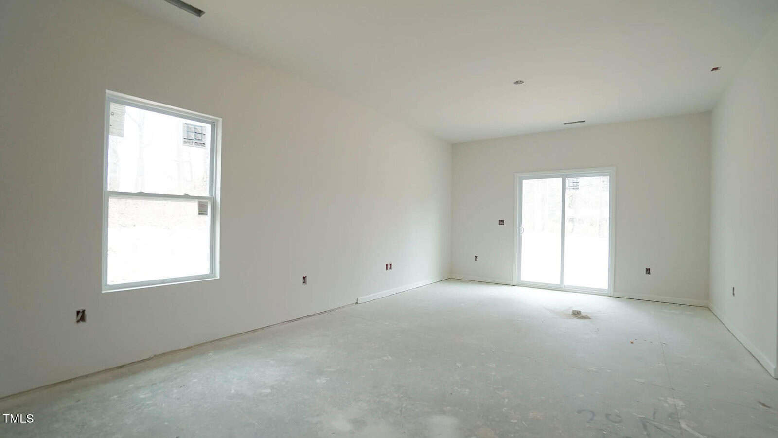 An empty room with unfinished floors, white walls, and a window on the left. There is a sliding glass door on the right leading outside, allowing natural light. The room is bare, with visible electrical outlets and ventilation openings.