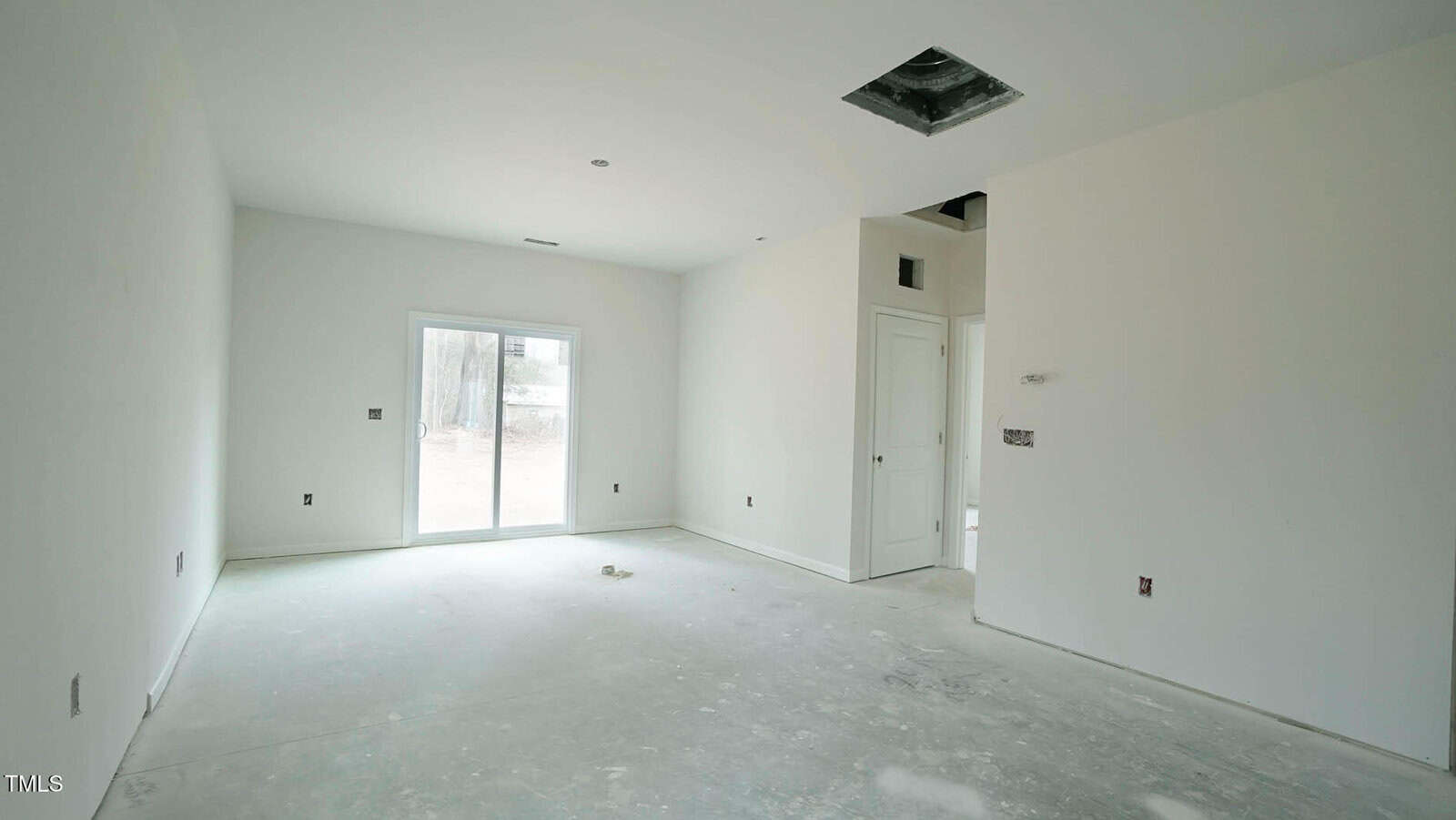 Empty room with unpainted walls and concrete floor. A sliding glass door on the left opens to the outside. A small square ceiling cutout is visible. An open doorway leads to another room on the right. Sparse light fixtures and outlets are installed.