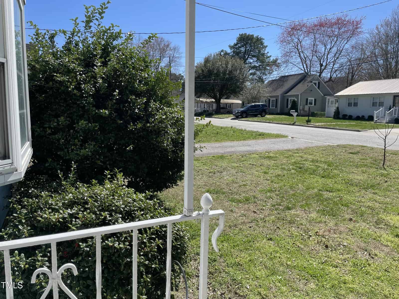 View from a porch showing a white railing, green bushes, a grassy yard, and a quiet residential street lined with houses and trees under a clear blue sky.