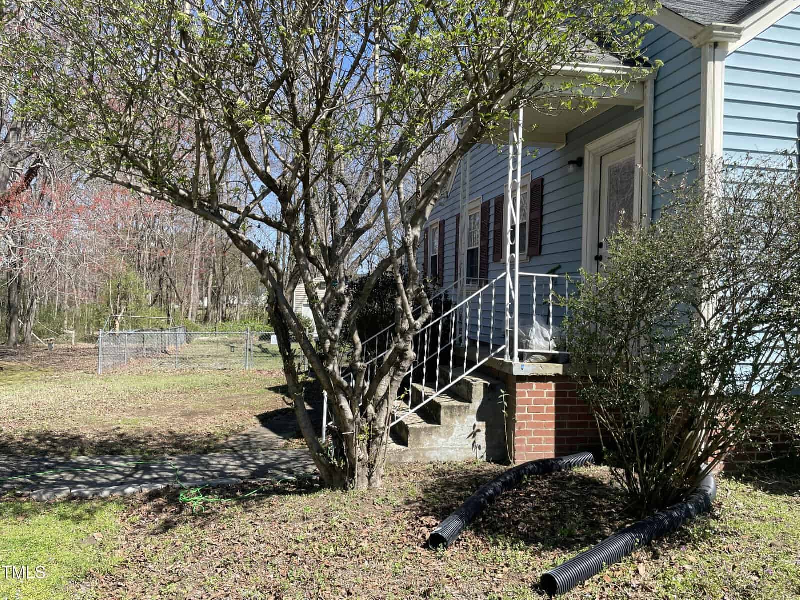A blue house with white trim and a small porch stands beside a large tree with budding leaves. Black drainage pipes extend from the foundation into a grassy yard on a sunny day.
