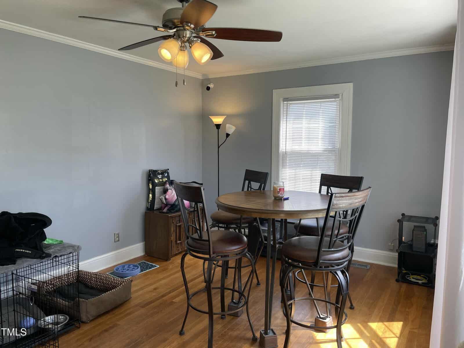 A small dining area with a round table and four chairs on a hardwood floor. The room has gray walls, a ceiling fan with lights, a standing lamp, a window with blinds, a dog crate, and pet supplies in the corner.