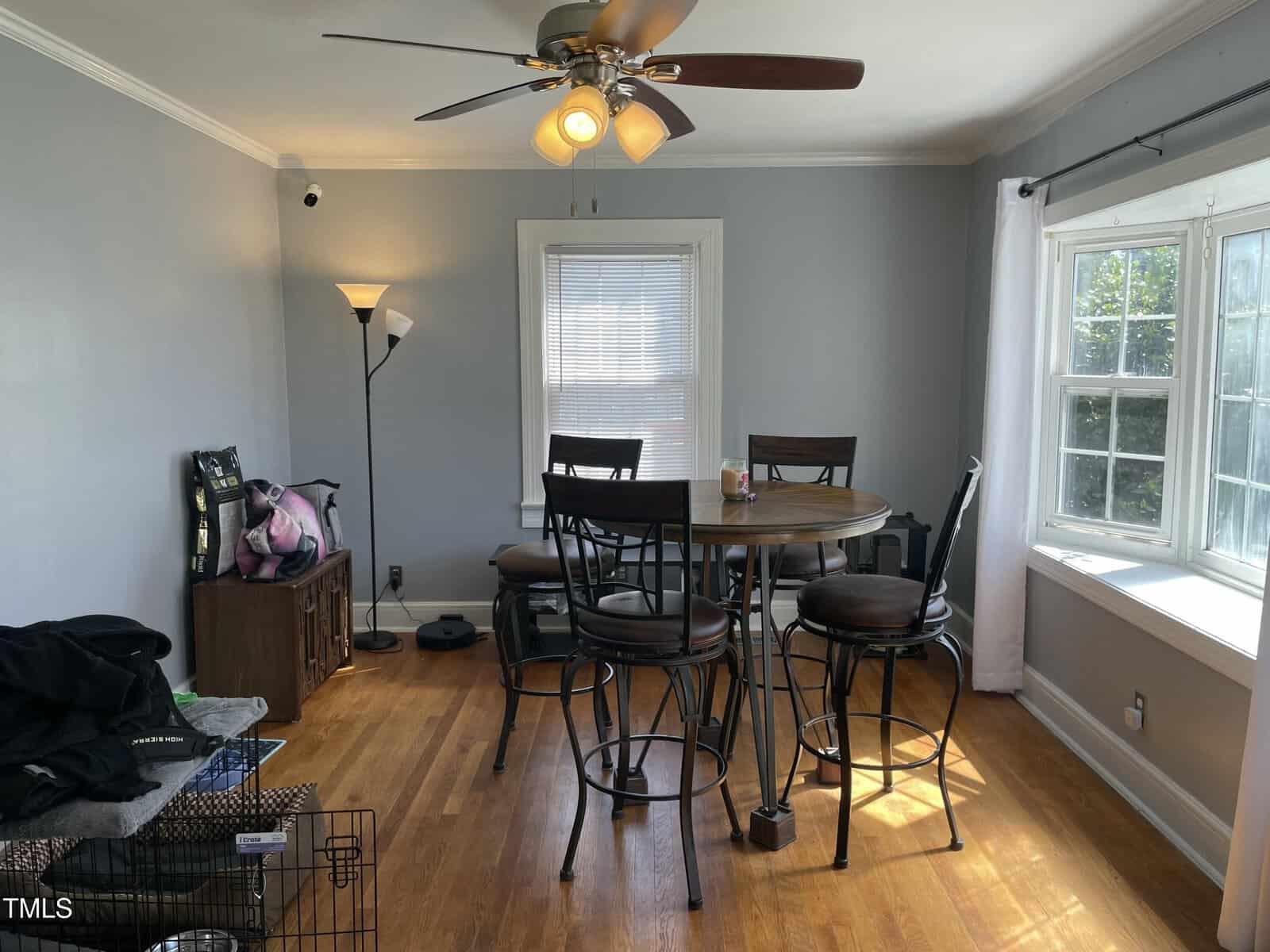 A small dining area with a round wooden table and four chairs on a hardwood floor, next to a large window. A ceiling fan with lights is on, and a floor lamp and miscellaneous items are in the corner.