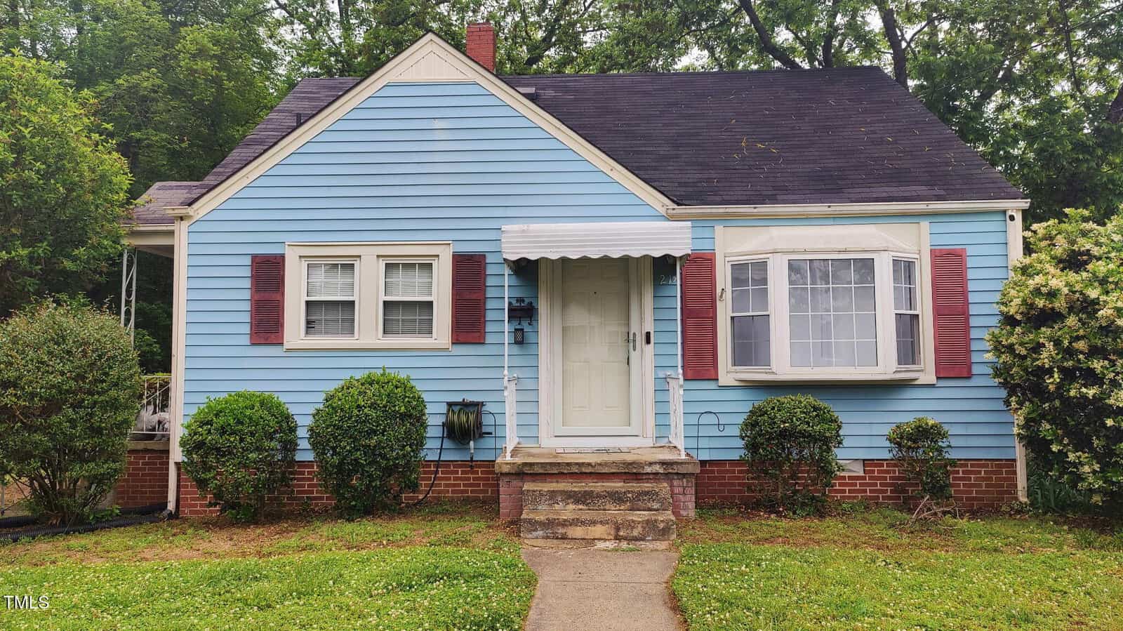 A small blue house with white trim at 214 Grace Street, Oxford, featuring maroon shutters, a white front door with an awning, brick accents at the base, lush bushes and shrubs in front, and trees in the background.