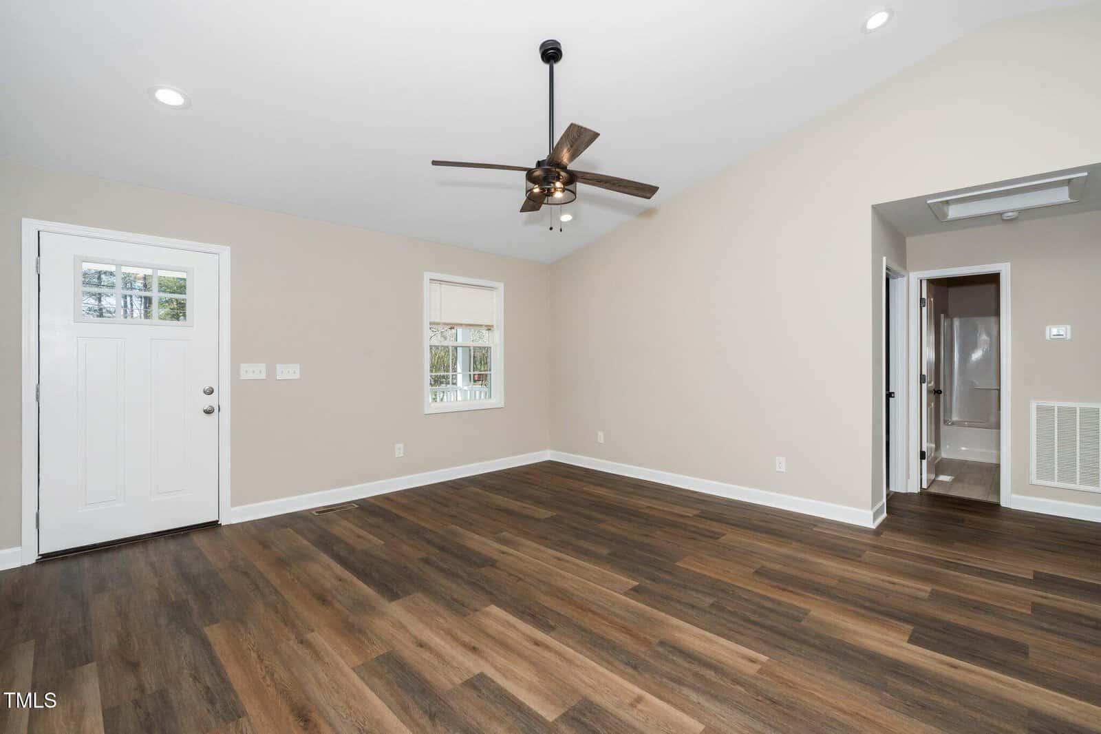 A vacant room with light beige walls and dark wood flooring. There is a ceiling fan with lights, a white entry door, a window, and an open doorway leading to a bathroom, visible on the right.