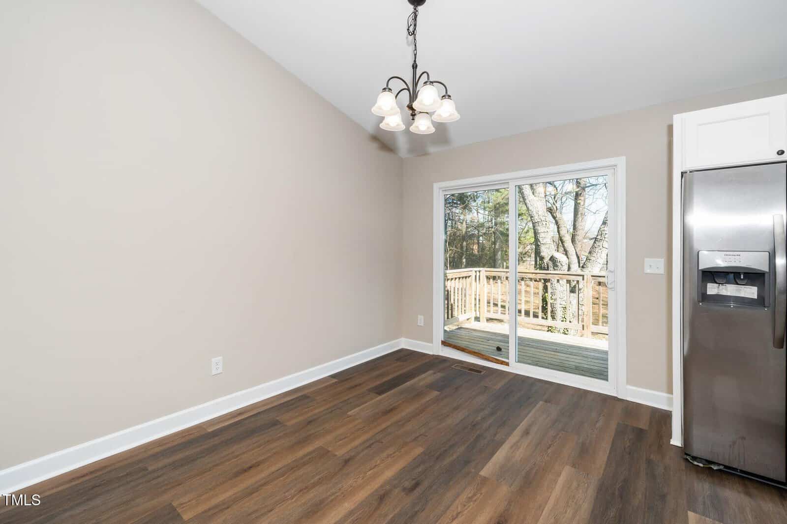 A room with wood flooring, a light fixture on the ceiling, and a sliding glass door leading to a wooden deck outside. A stainless steel refrigerator is on the right, and the wall is painted light beige. Trees are visible through the door.