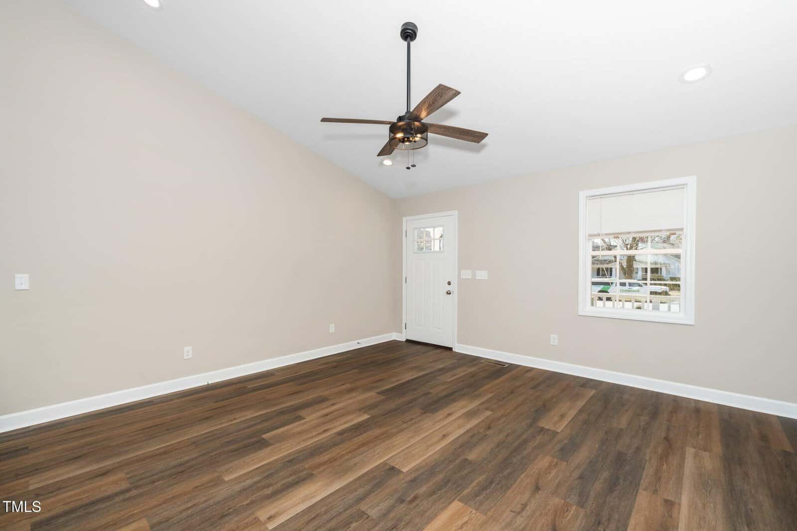 An empty room with light-colored walls and wood flooring. The room has a vaulted ceiling with a dark ceiling fan. A white framed window and a white door with a window are on one wall. Recessed lighting is installed in the ceiling.
