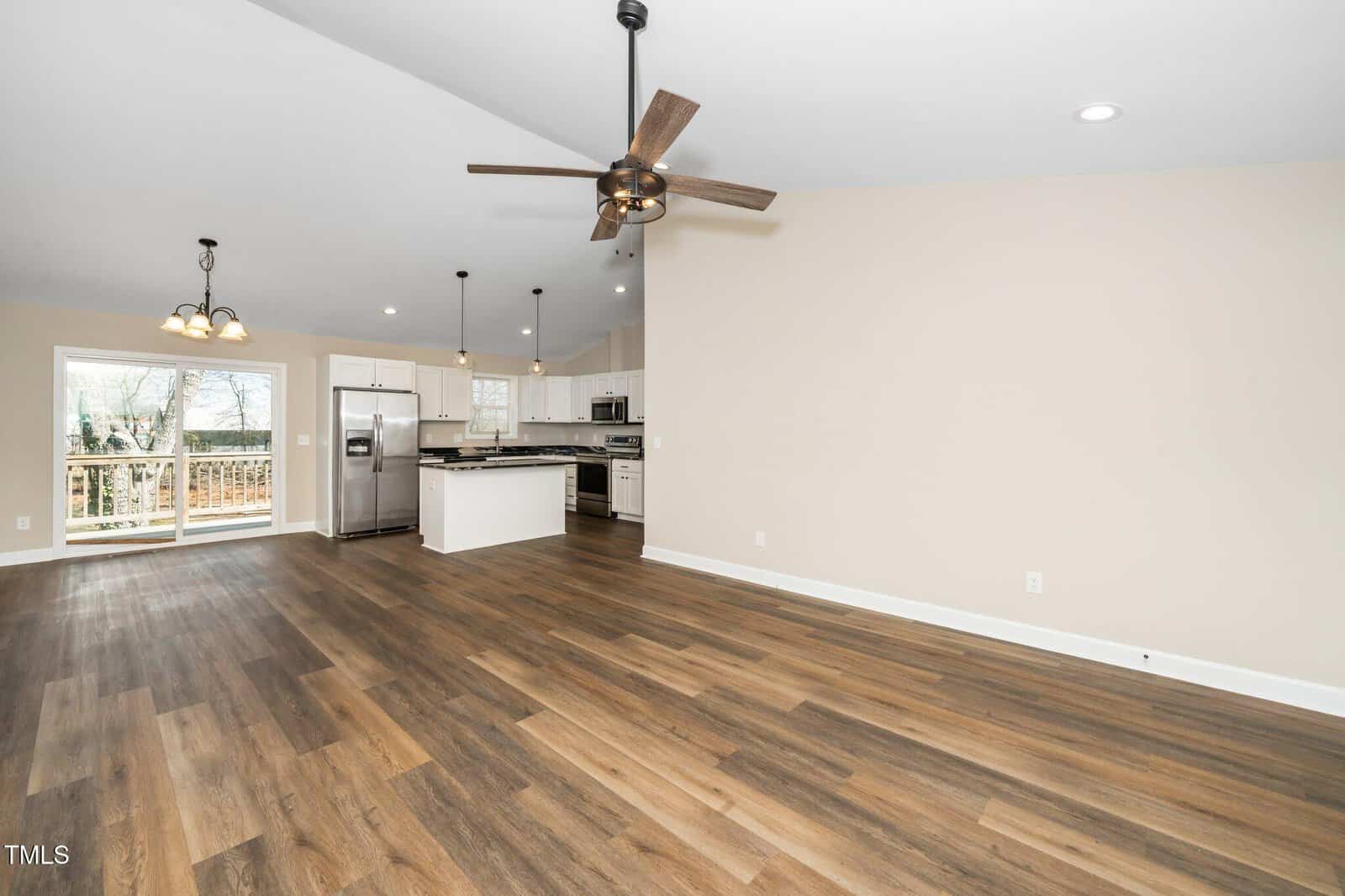 Open-concept kitchen and living area with wood flooring. The kitchen features white cabinets, a dark countertop, and stainless steel appliances. A ceiling fan is in the living area, and sliding glass doors open to a deck with a view of trees.