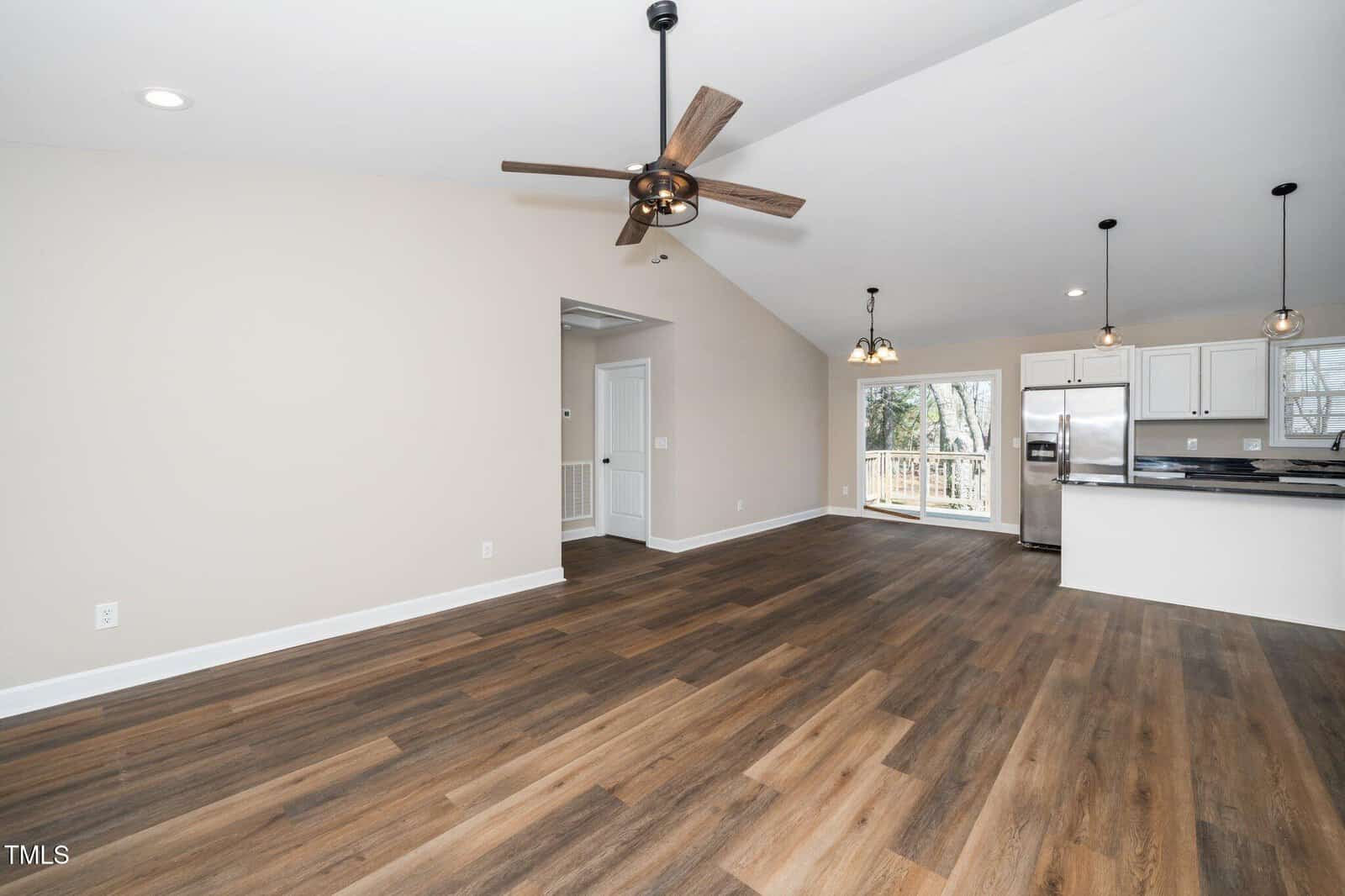 Spacious open-concept interior featuring a living area with a ceiling fan, wood-style flooring, and high ceilings. In the background, a modern kitchen includes a breakfast bar, pendant lights, and stainless steel appliances. Large sliding door to a deck.