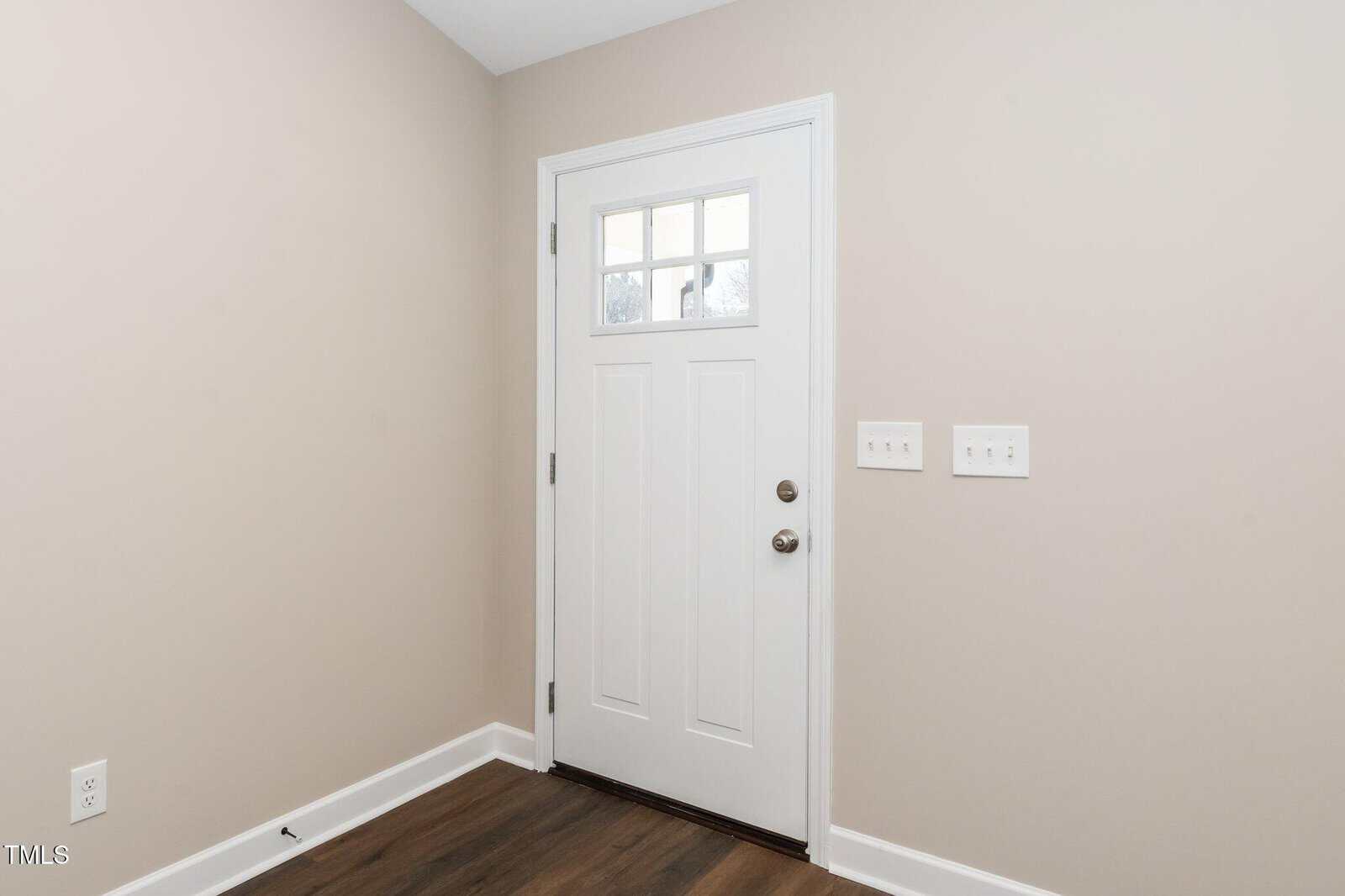 A white front door with a small window panel is set in a beige wall. The door has a silver doorknob and deadbolt. Two light switches are on the wall to the right. The floor is wooden.