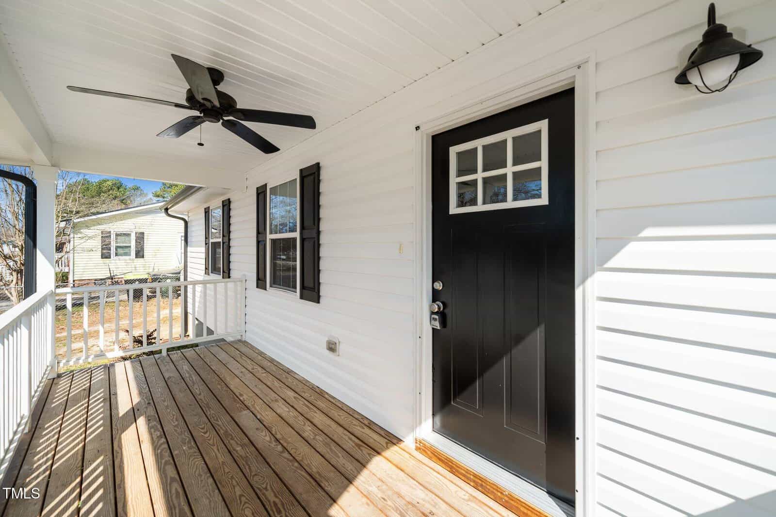 A wooden porch features a black door with a window panel and a metal handle. A ceiling fan and light fixture are mounted on the white ceiling. The siding is white with black shutters. A white railing borders the porch, and a neighboring house is visible.