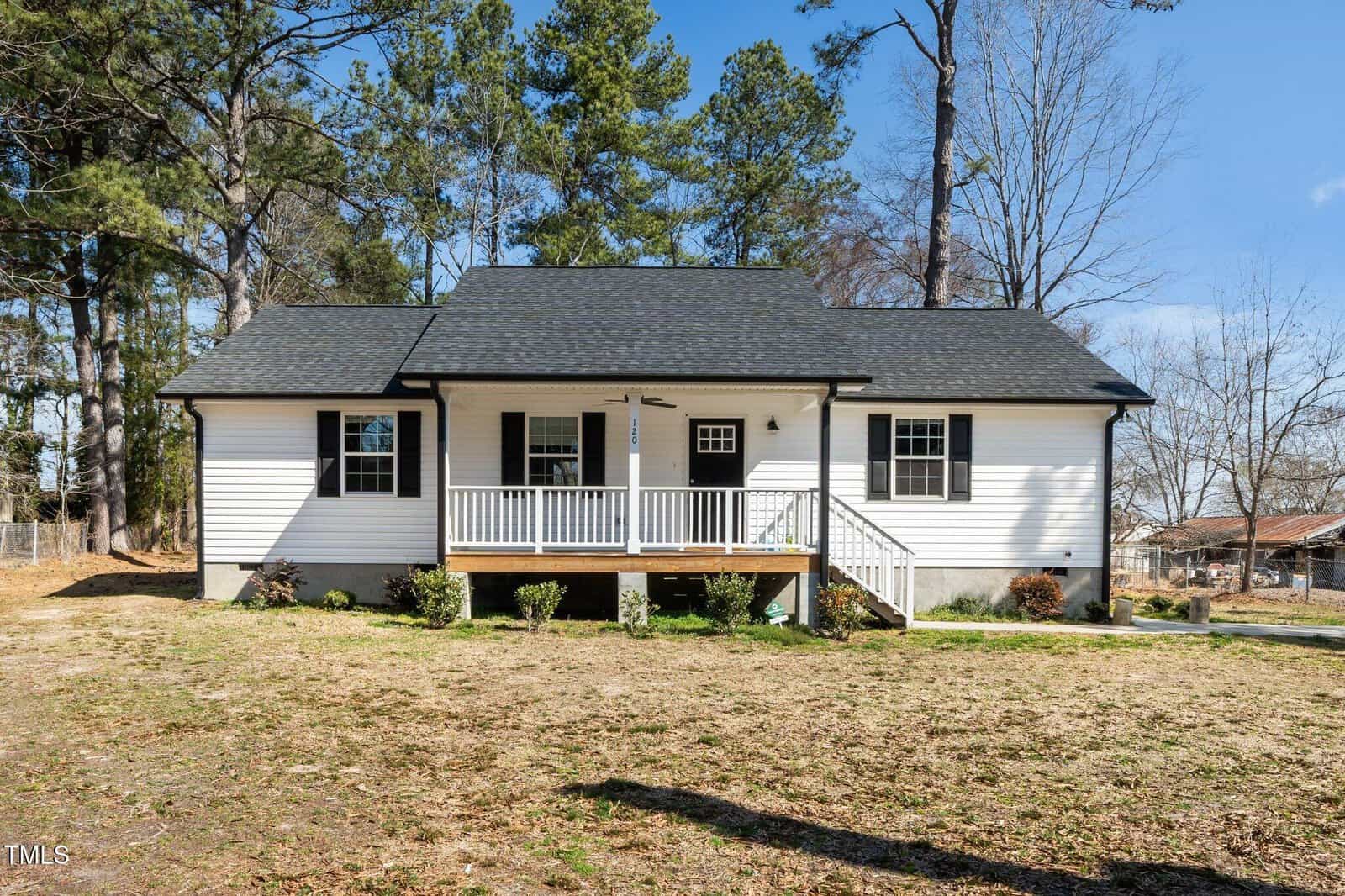 A charming, single-story white house with a blue roof and black shutters sits serenely at 120 W College Street in Oxford. It features a front porch with white railings, steps leading to the door, a spacious grassy yard, and tall trees surrounding the property.