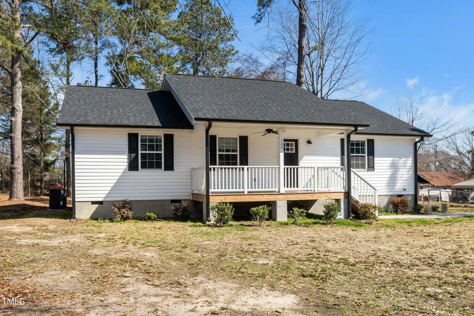 A white single-story house with a black roof and a small front porch. The porch has a ceiling fan and is supported by four posts. The yard has sparse grass, and there are trees in the background under a clear blue sky.