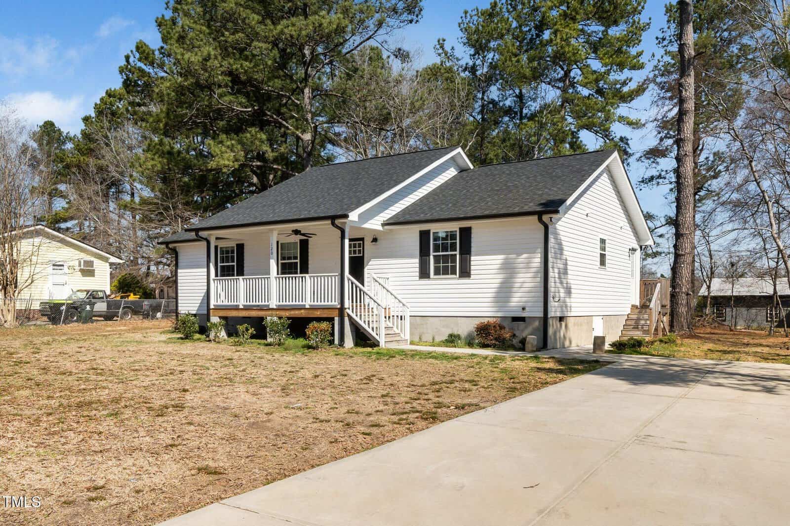 A single-story white house with a dark roof is surrounded by a few trees. It features a small front porch with railing. The yard is mostly grass with some bare patches, and there is a wide concrete driveway leading to the house.