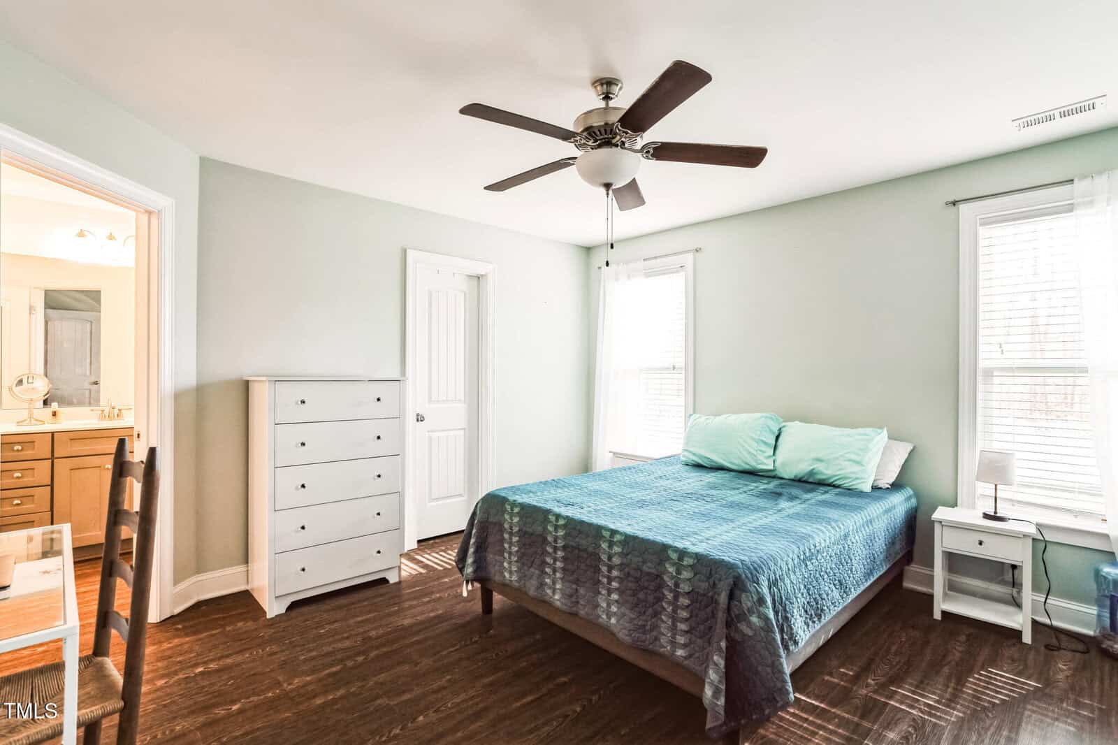 A bedroom with a double bed covered in a blue patterned quilt, two pillows, and a ceiling fan above. On the left, there's a chest of drawers and a partial view of a chair and bathroom. Two windows allow natural light in. The floor is dark wood.