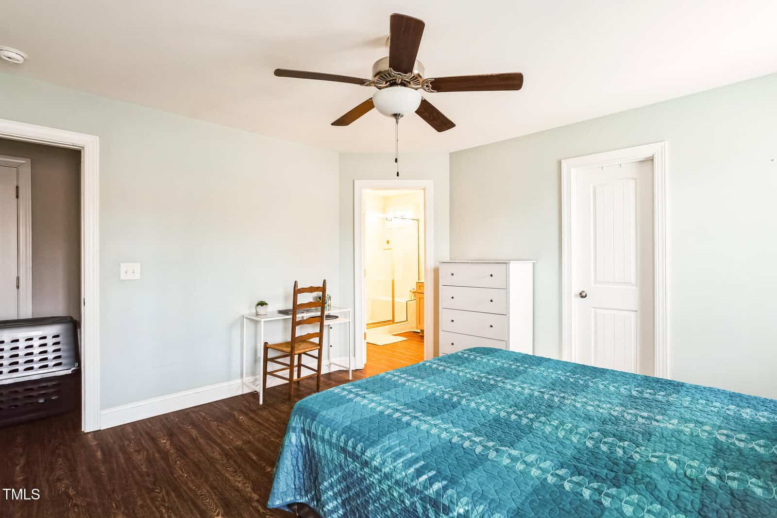 Bedroom with a teal bedspread, wooden chair, and small table. Light green walls and dark wood flooring. Ceiling fan above. Doorways lead to a bathroom and a closed room. White dresser against the wall. Bright bathroom with shower visible.