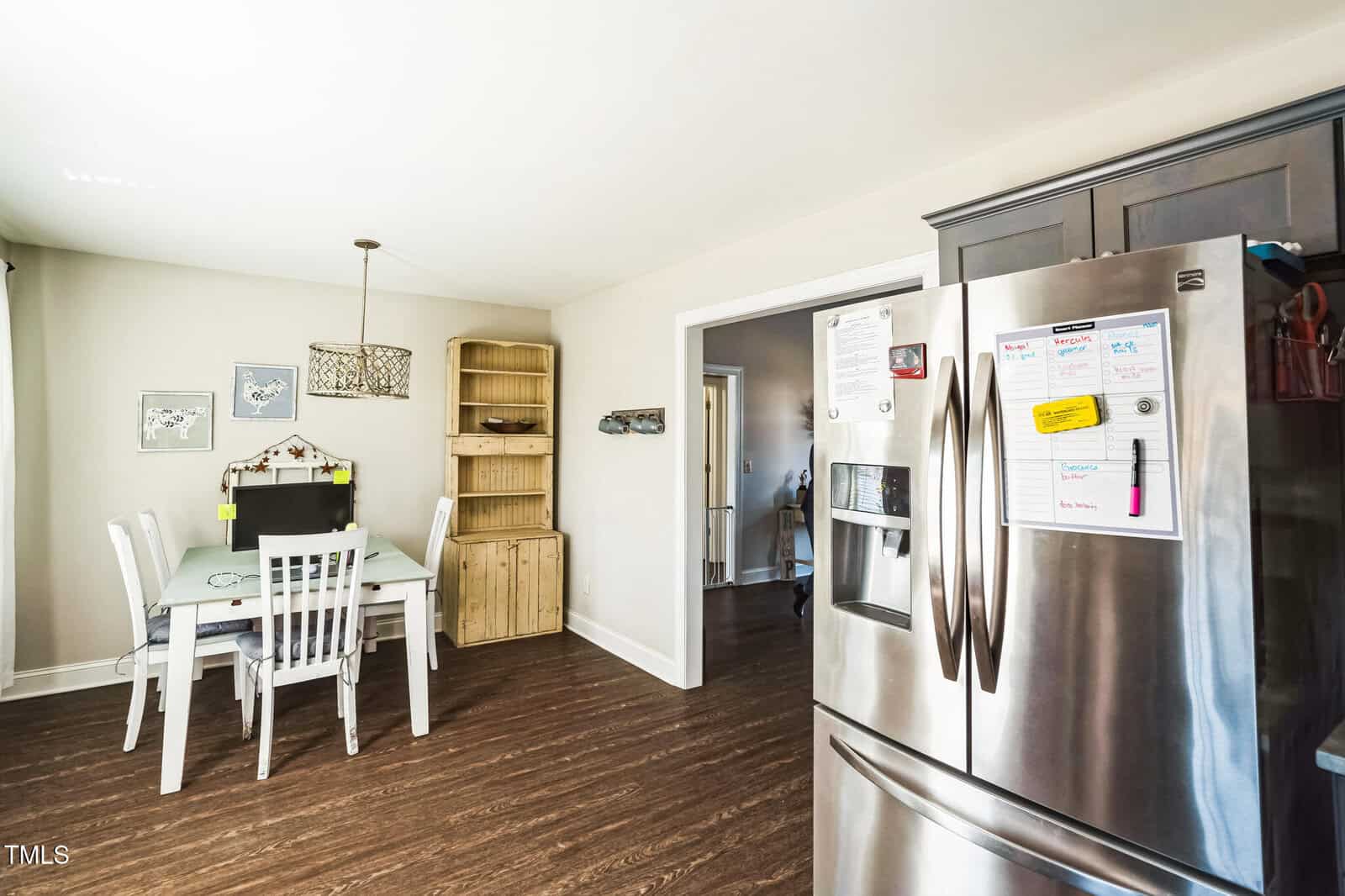 A kitchen with dark wooden floors, a refrigerator with notes attached, and a light-colored wooden dining table with four white chairs. A wooden shelving unit stands in the corner. A window on the left brings in natural light.