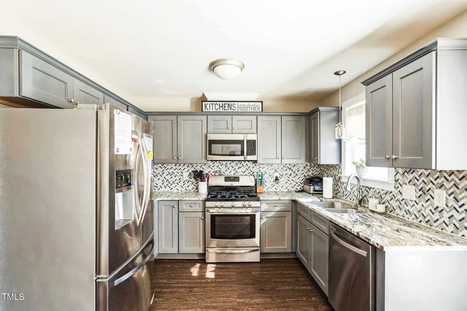 A kitchen with gray cabinets, stainless steel appliances, and a herringbone-patterned backsplash. There is a refrigerator, stove, microwave, and dishwasher. The countertop is granite, and a window above the sink lets in natural light.