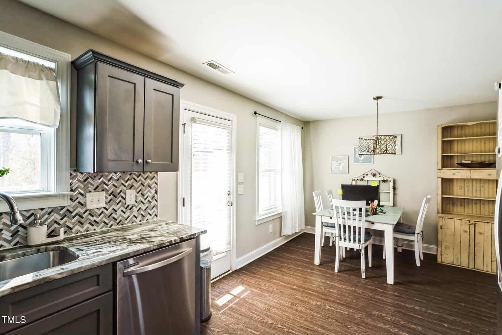 A kitchen and dining area with gray cabinets, a marble countertop, and a herringbone backsplash. The dining area has a table with white chairs under a geometric chandelier. A wooden cabinet stands against the wall. Sunlight enters through windows and a door.