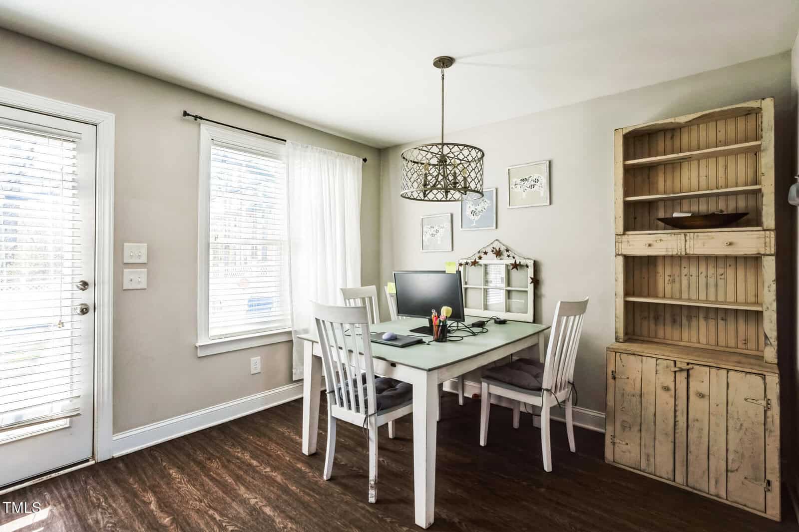 A dining area features a white rectangular table with three white chairs. A computer monitor and keyboard are on the table. A decorative light fixture hangs above. A wooden hutch stands against one wall. Two windows with blinds provide natural light.