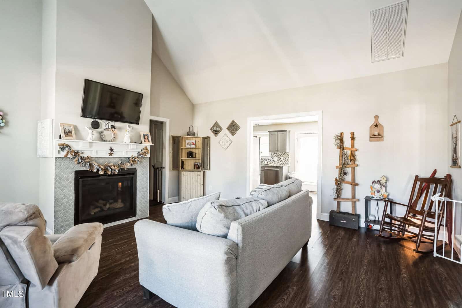 A living room with a high ceiling features a gray sofa and armchair on a dark wood floor. A TV is mounted above a fireplace decorated with ornaments. Shelves with decor are on either side, and light enters from an adjacent room with kitchen view.