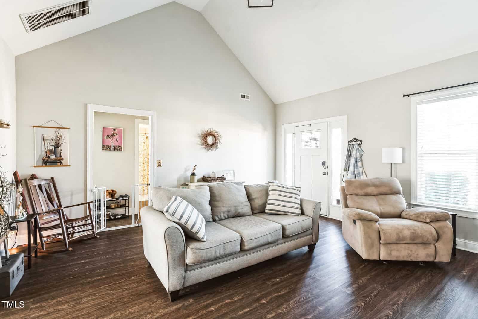 Living room with vaulted ceiling, featuring a beige sofa with striped pillows, a beige recliner, and decorative elements like a wall wreath and rocking chair. A large window with blinds allows natural light. Dark hardwood flooring throughout.
