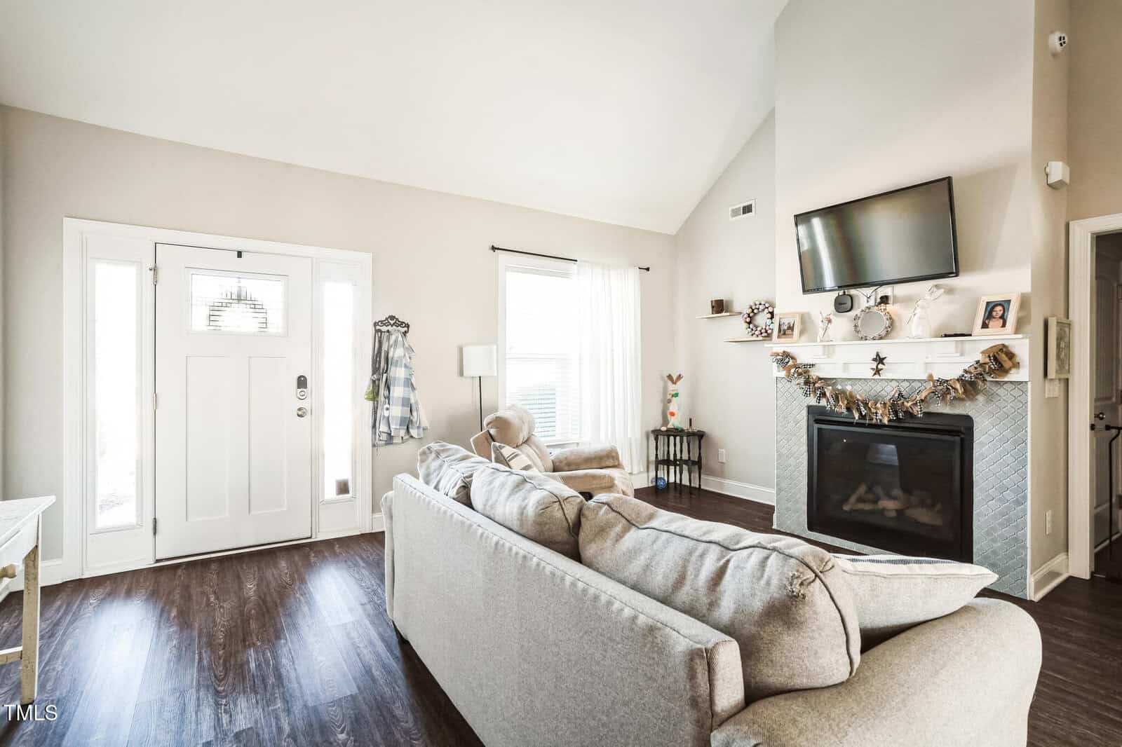 A living room with a gray couch, a wall-mounted TV above a fireplace, and a small round side table. The room has wooden flooring, a high ceiling, and large windows with white curtains. A white front door with a window is on the left.