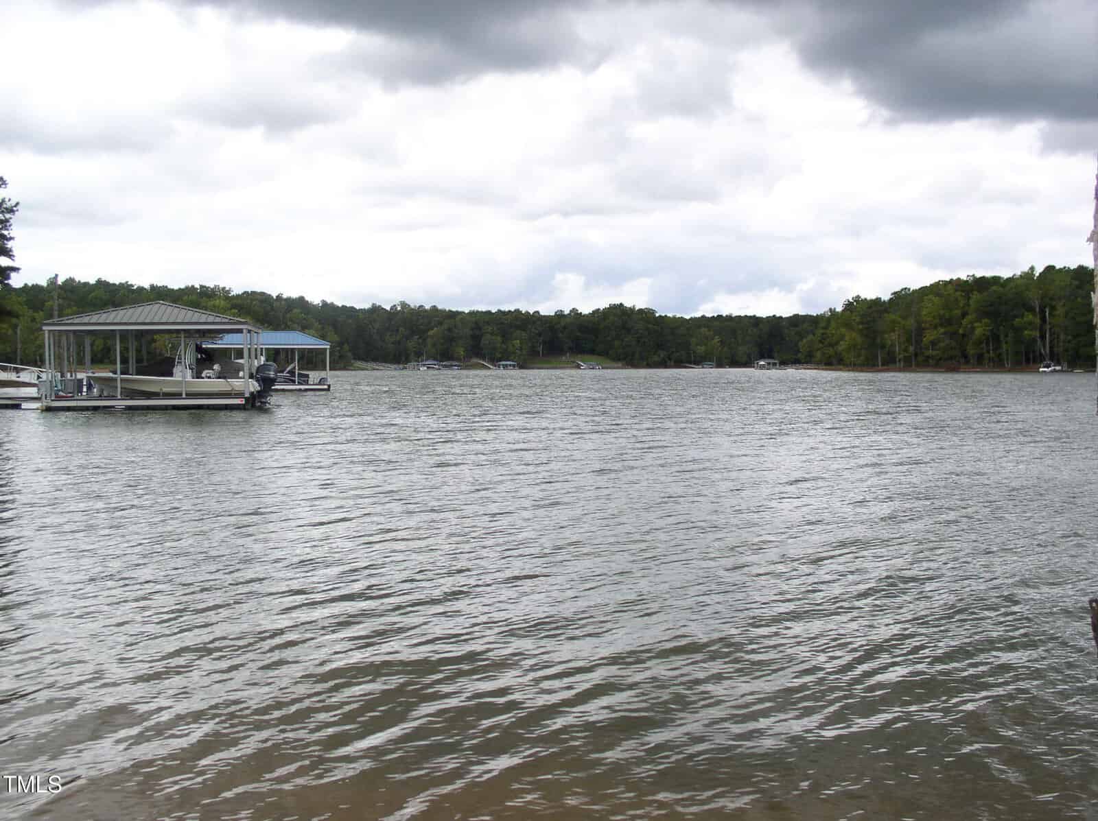 A large body of water with a gentle wave texture under a cloudy sky. A boat is docked under a covered structure on the left side. Trees line the far shore in the background, creating a forested backdrop.