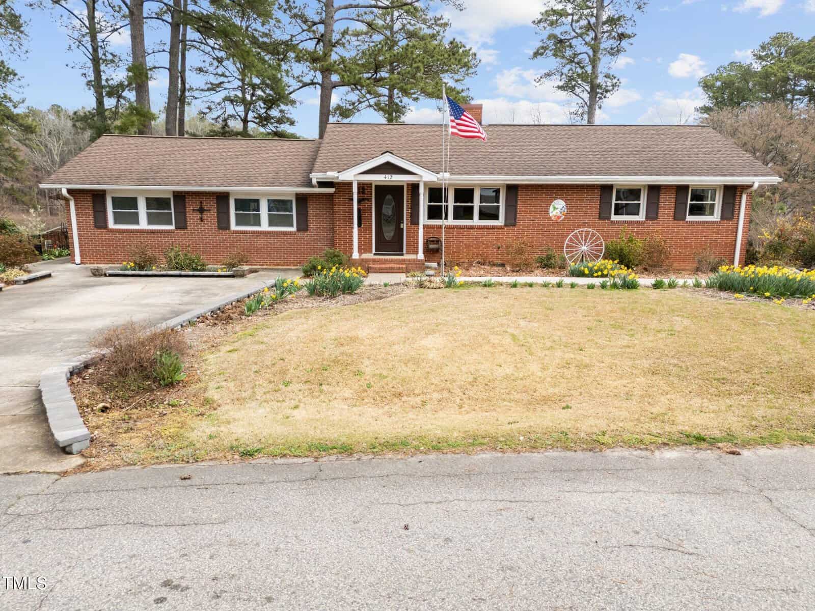 A single-story red brick house with a gabled roof, white trim, and an American flag on a pole in the front yard. There are flower beds, a decorative wagon wheel, and a concrete driveway and walkway.