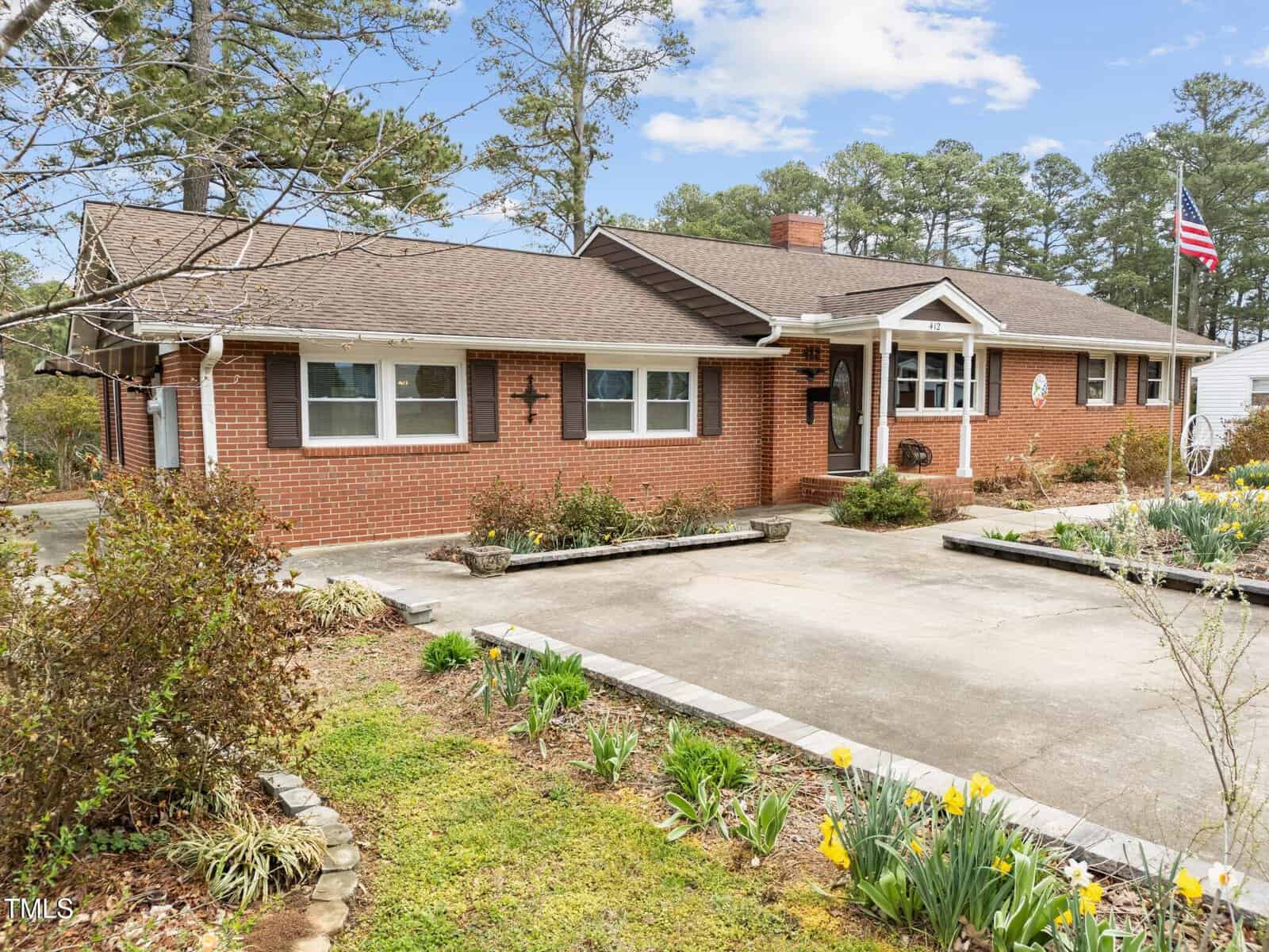 Single-story red brick house with white trim, gabled roof, a front porch, and flower beds with blooming yellow flowers. Concrete driveway in front, American flag on the right side, and tall trees in the background.
