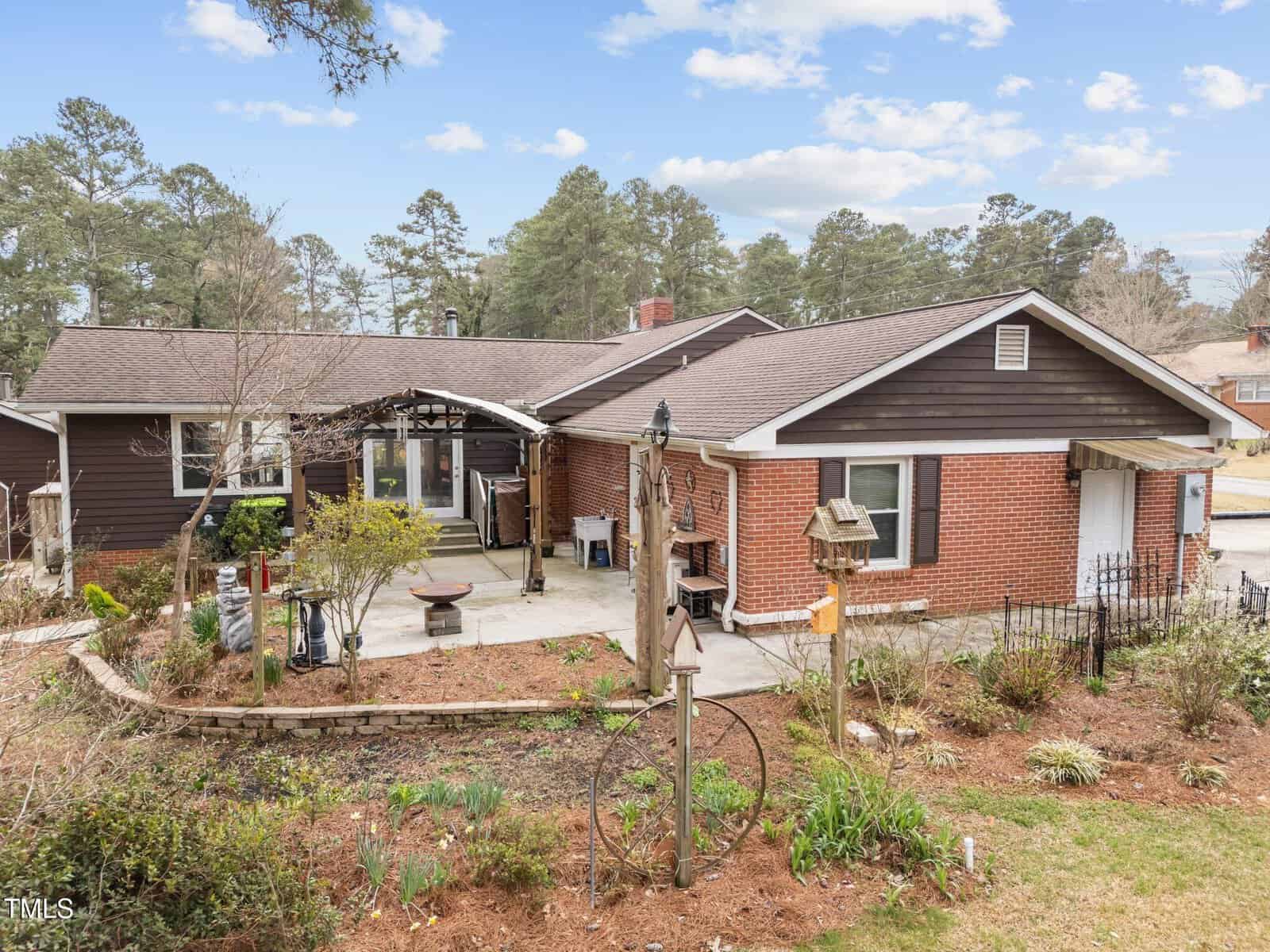 One-story house with red brick and dark brown siding, surrounded by a garden with bare and green plants. Concrete patio area in front. Decorative birdhouse and metal garden art visible. Trees and blue sky in the background.