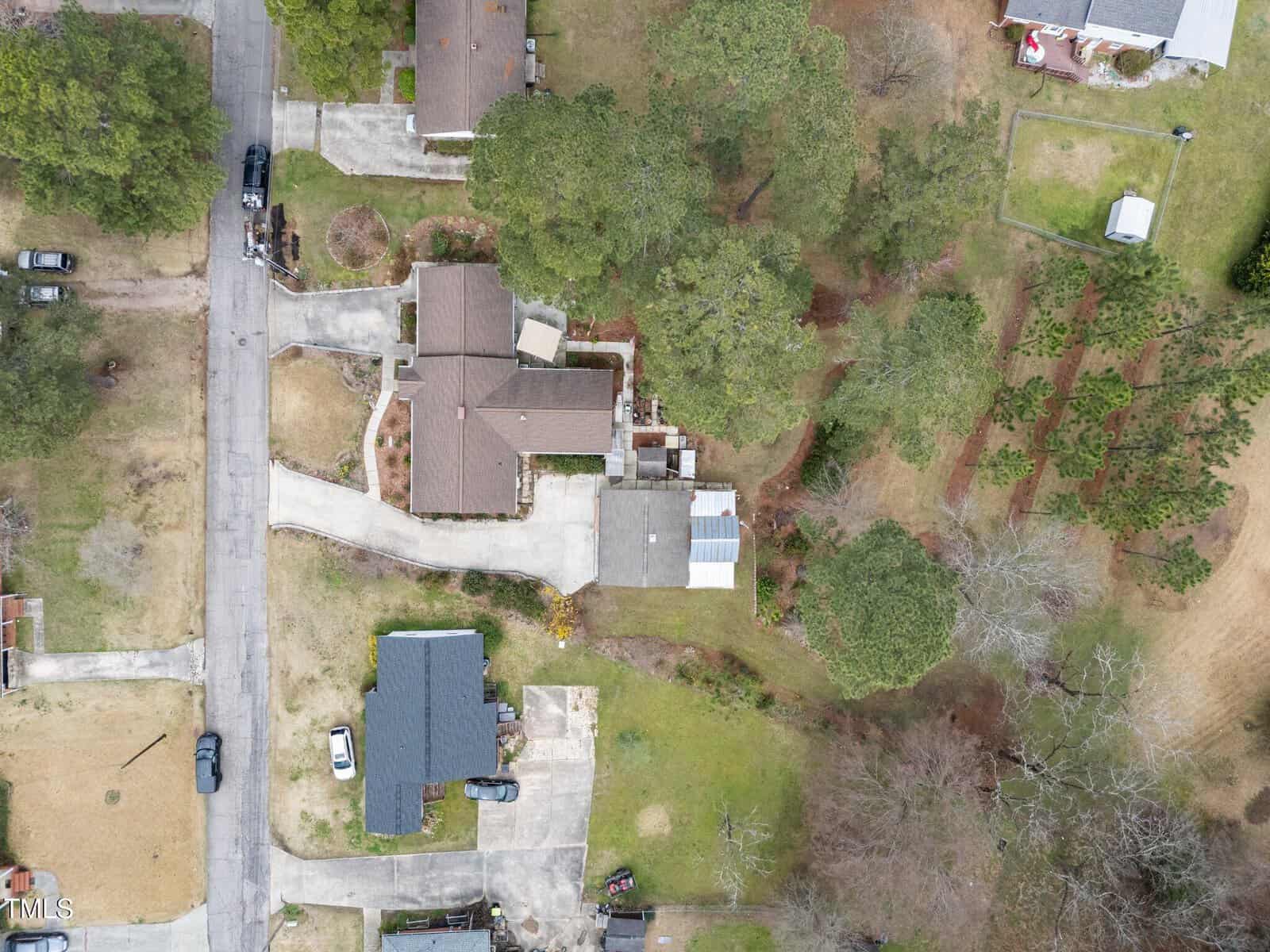 Aerial view of a suburban neighborhood showing several houses with driveways, cars parked on the street and driveways, trees, grassy lawns, and a garden area in the upper right section of the image.