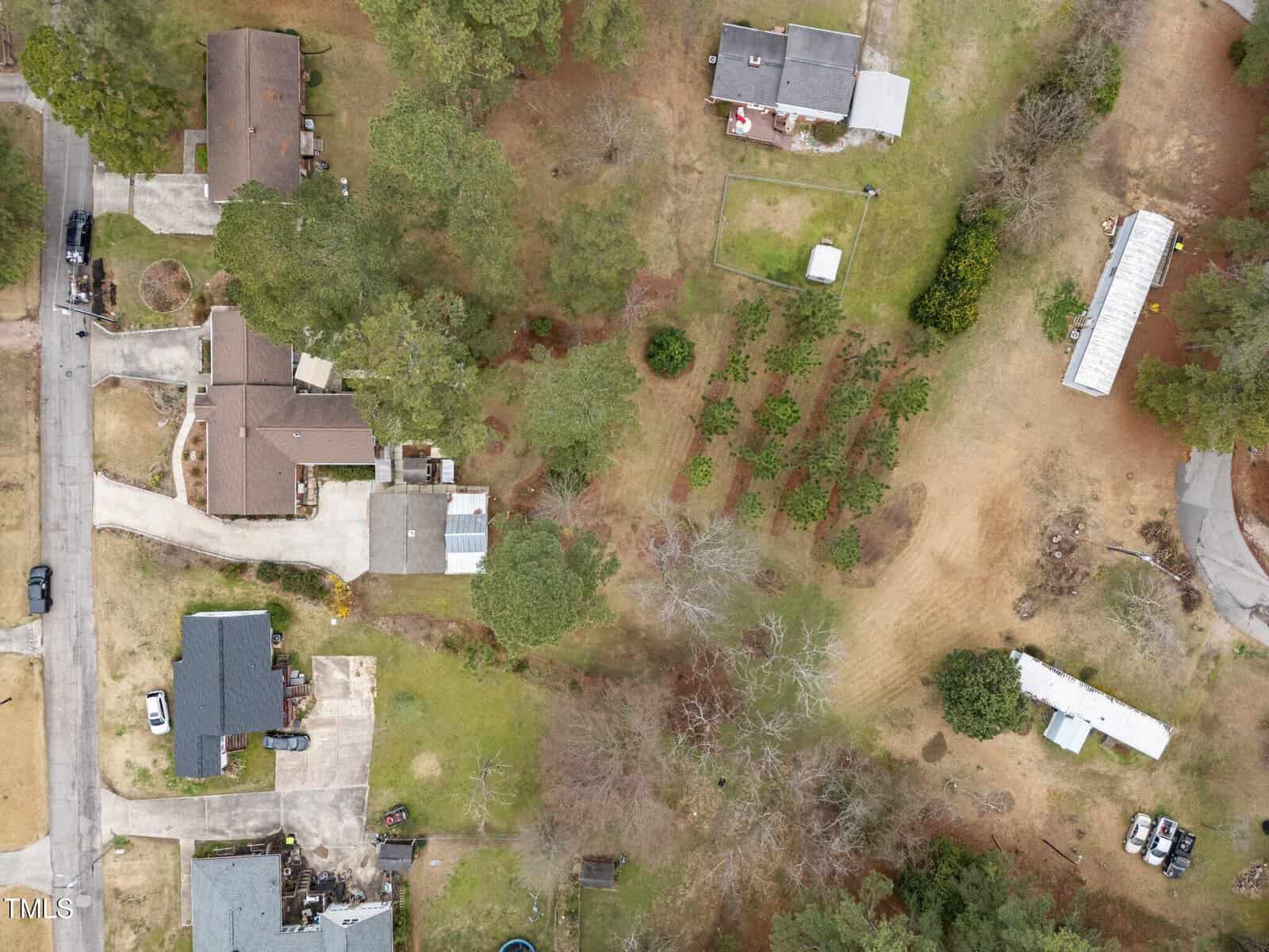 Aerial view of a suburban neighborhood showing several houses, driveways, trees, grassy areas, and a few outbuildings. The landscape includes both open and wooded spaces with some vehicles parked near the homes.