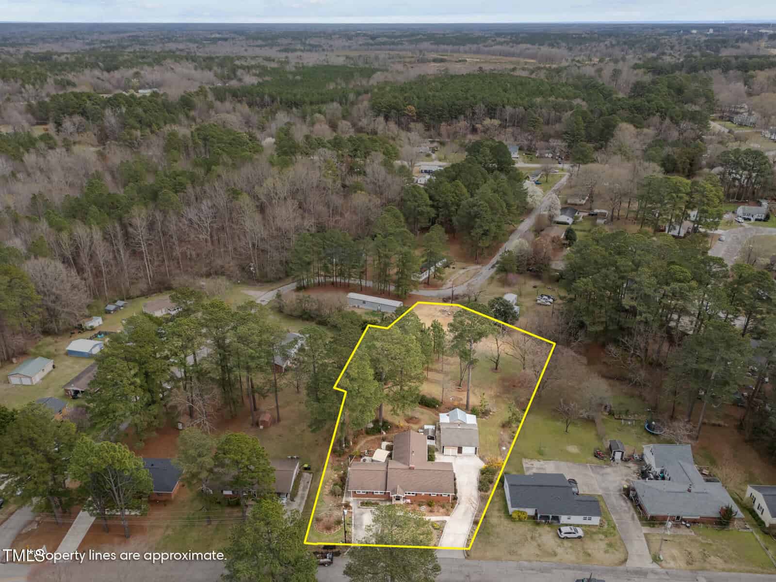 Aerial view of a residential property outlined in yellow, showing a large brick house, driveway, and surrounding trees in a suburban neighborhood. The caption notes the property lines are approximate.