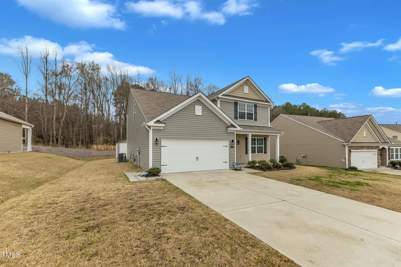 A two-story house with gray siding and white trim, featuring an attached two-car garage. The home is set on a sloping lawn, bordered by trees in the background under a blue sky with clouds.