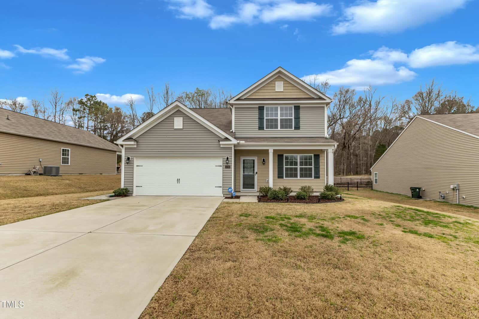 A two-story house with gray siding and a gabled roof. The house has a double garage and a small front yard with shrubs. The driveway and lawn are neatly maintained. It's a clear day with a blue sky and scattered clouds.
