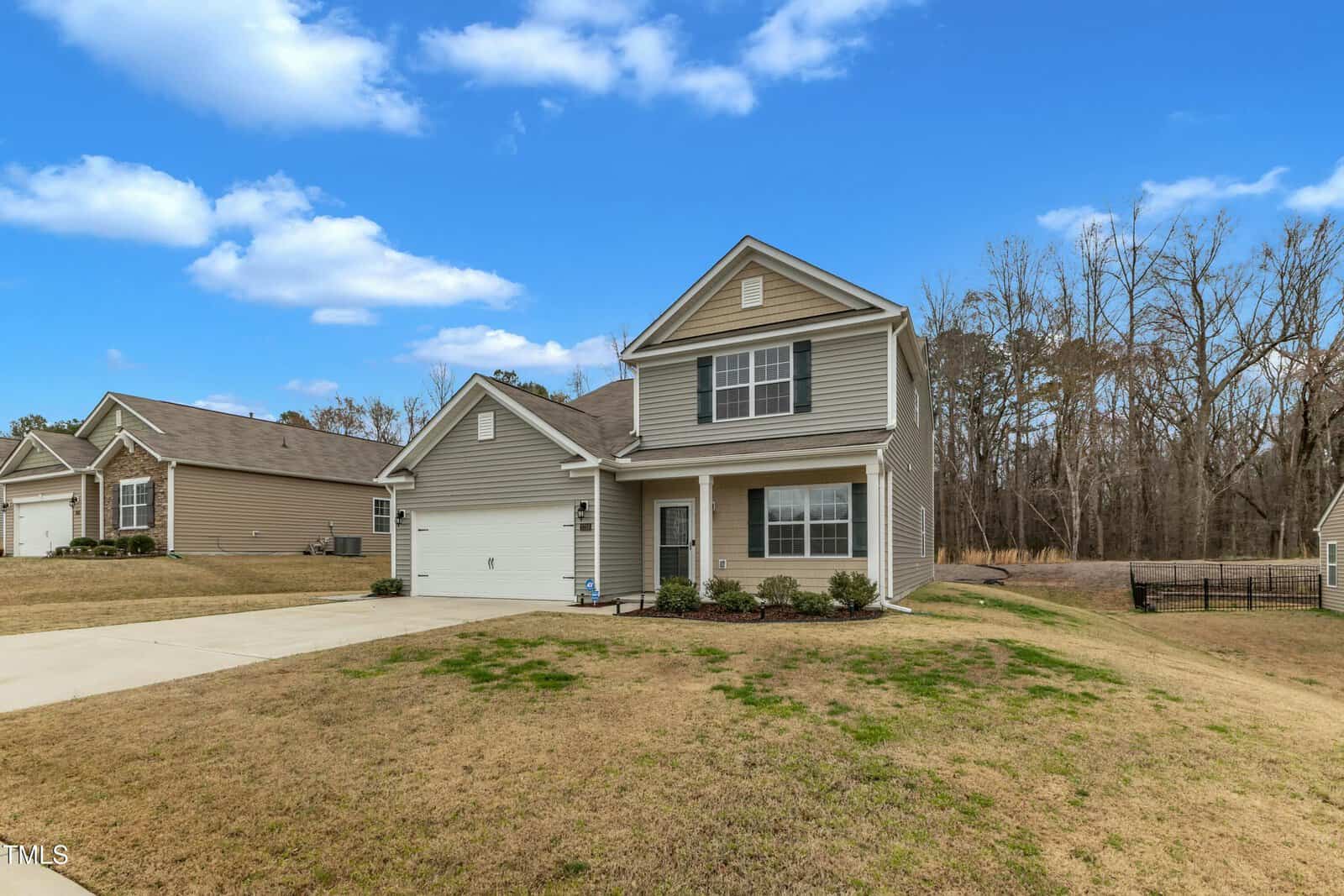 A two-story house with gray siding and white trim sits on a grassy lawn. It has a covered front porch, a driveway leading to a two-car garage, and borders a dense stand of trees. The sky is clear with a few clouds.