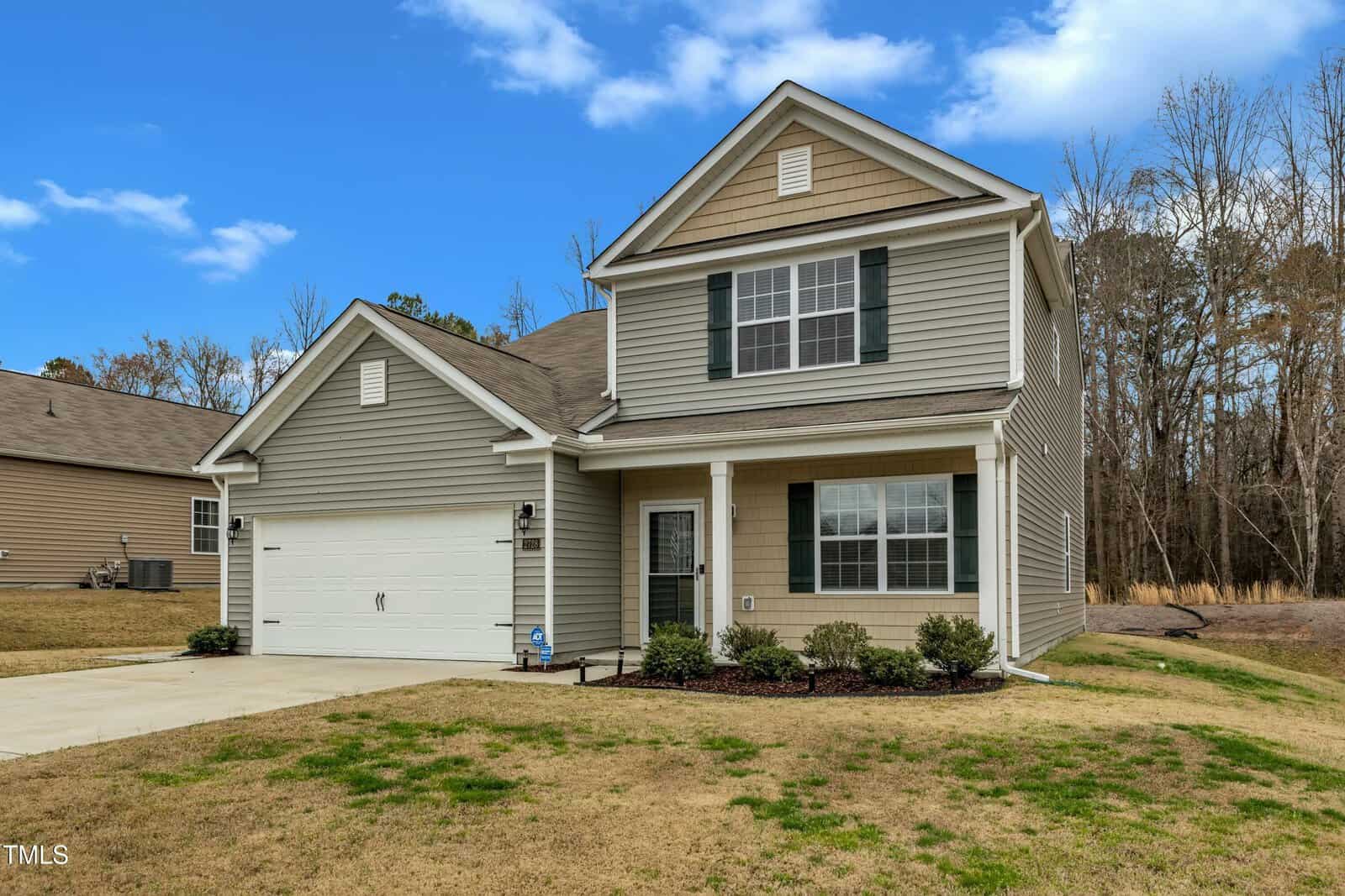 A two-story house with gray siding and white trim, featuring a front porch and a double garage. The property has a small lawn and is set against a backdrop of trees. The sky is partly cloudy.