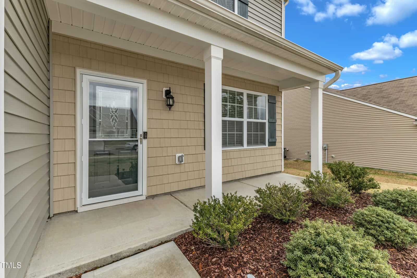 The image shows the exterior of a house featuring a small front porch with two white columns. A glass storm door and window with blinds are present. The siding is beige with white trim. Shrubs line the mulched area beside the porch. The sky is partly cloudy.