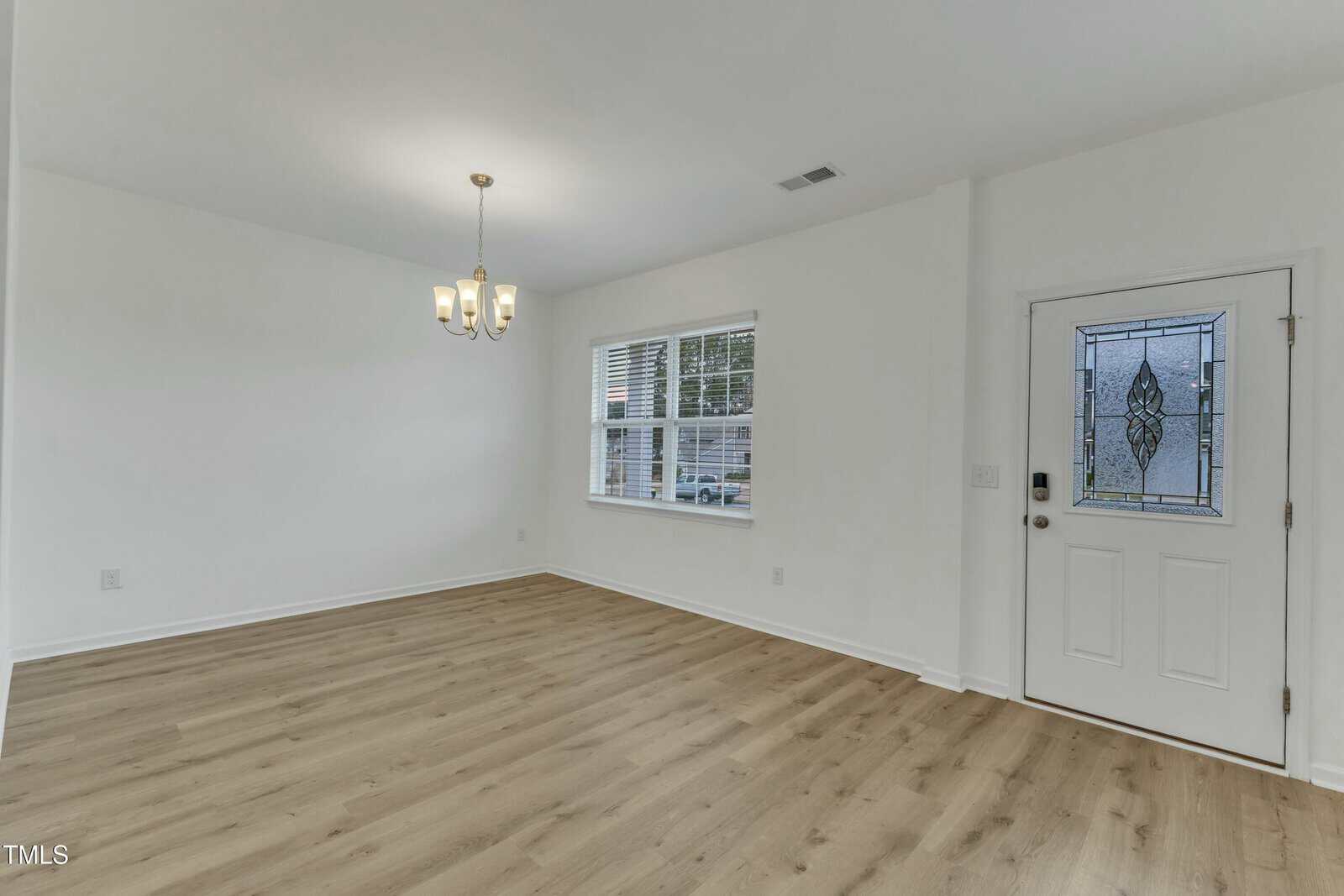 An empty room with light wooden flooring and white walls. There is a window with blinds on the left wall and a decorative glass-paneled white door on the right. A ceiling light fixture hangs in the center, and sunlight is visible through the window.
