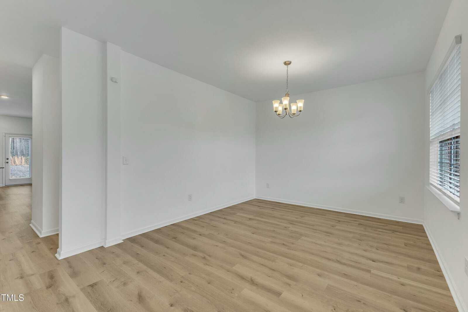 An empty room with light wood flooring and white walls. A chandelier hangs from the ceiling, and there are windows on the right side. A hallway is visible to the left, leading to another room with a door.