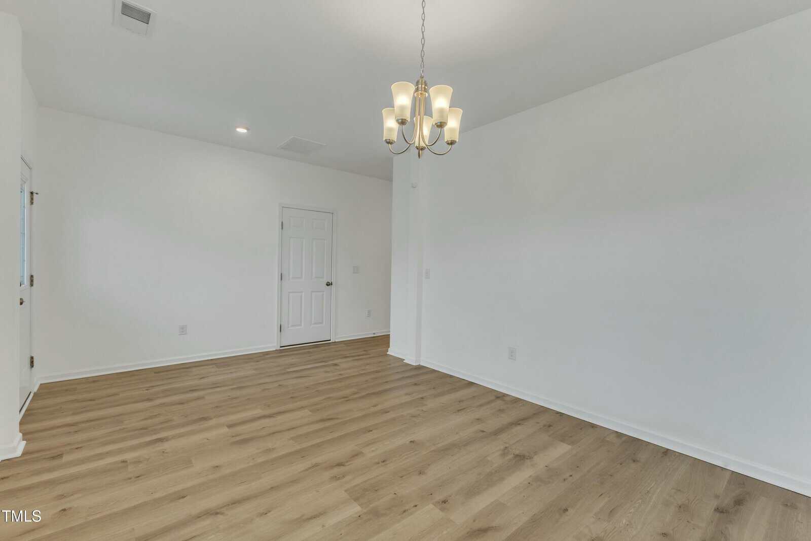 An empty room with light-colored walls and wood flooring. A ceiling light fixture with multiple bulbs hangs from above. A closed door is visible on the far wall, with another partially visible door on the left.