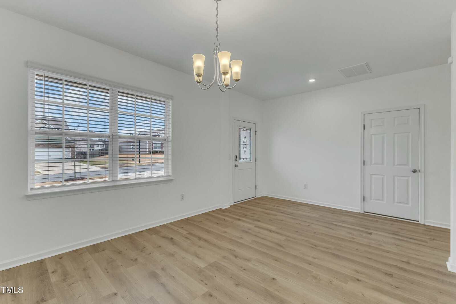 A bright, empty room with light colored wood flooring and white walls. A window with blinds is on the left wall. A door is in the background next to another closed door. A ceiling light fixture with five bulbs is hanging above.