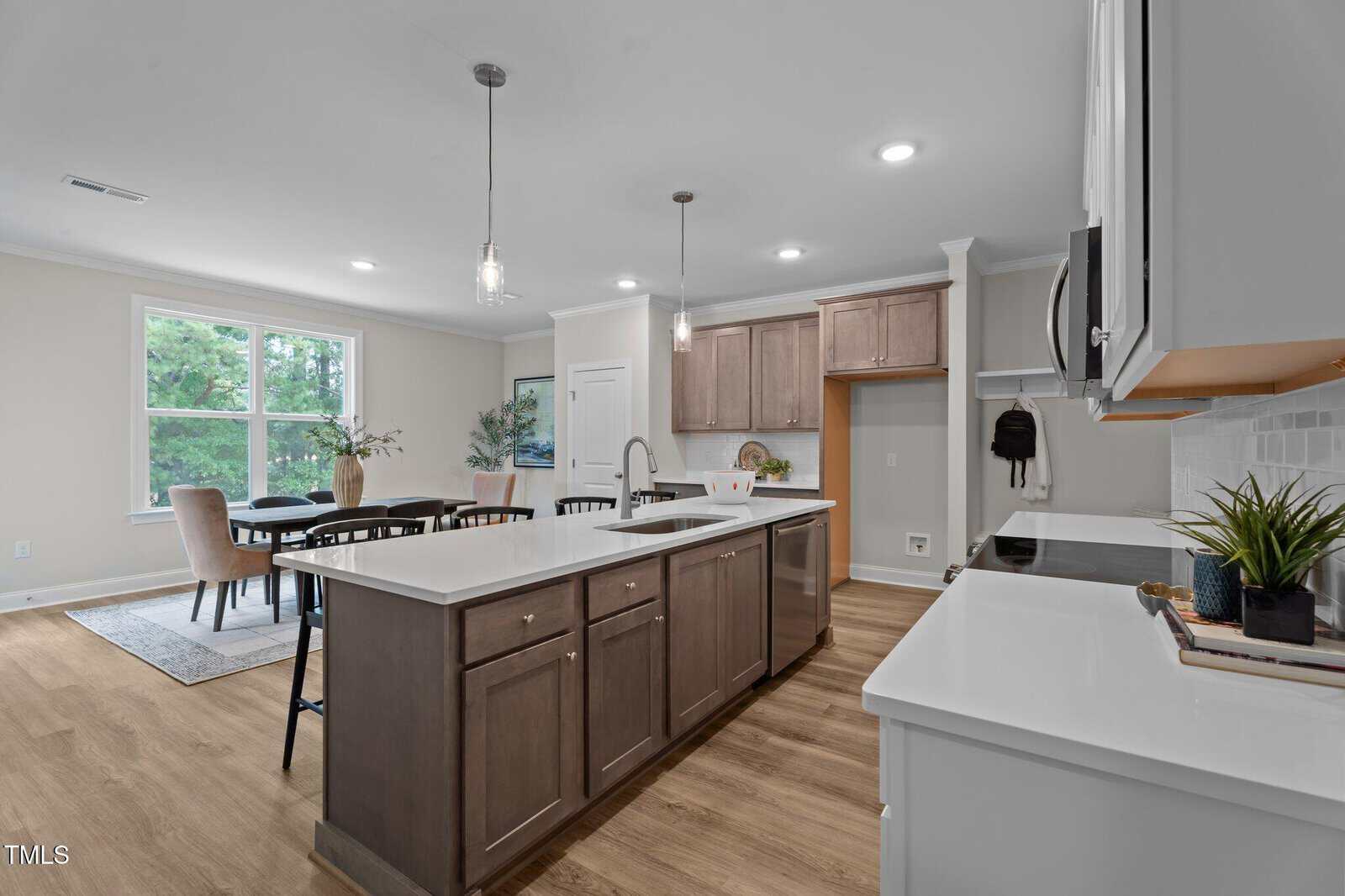 Modern kitchen with a large island, white countertops, wooden cabinets, stainless steel appliances, and wood flooring. A dining area with a table and chairs is visible near a window overlooking greenery.