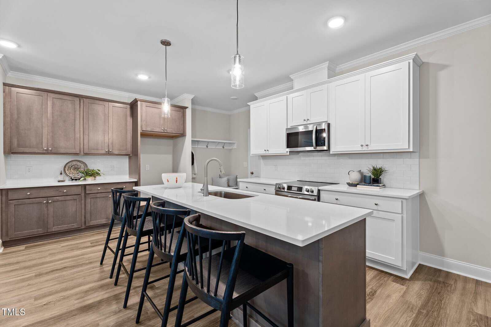 Modern kitchen with white cabinets, stainless steel appliances, a large island with sink, four black barstools, wood flooring, and pendant lights. A separate area features brown cabinets and decorative items.