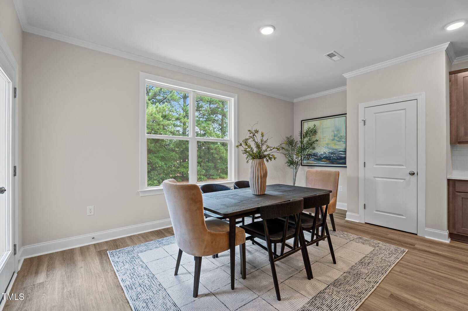 A dining area with a black rectangular table, four beige chairs, a vase with greenery, a large window with trees outside, light walls, wood flooring, and a framed painting on the wall.