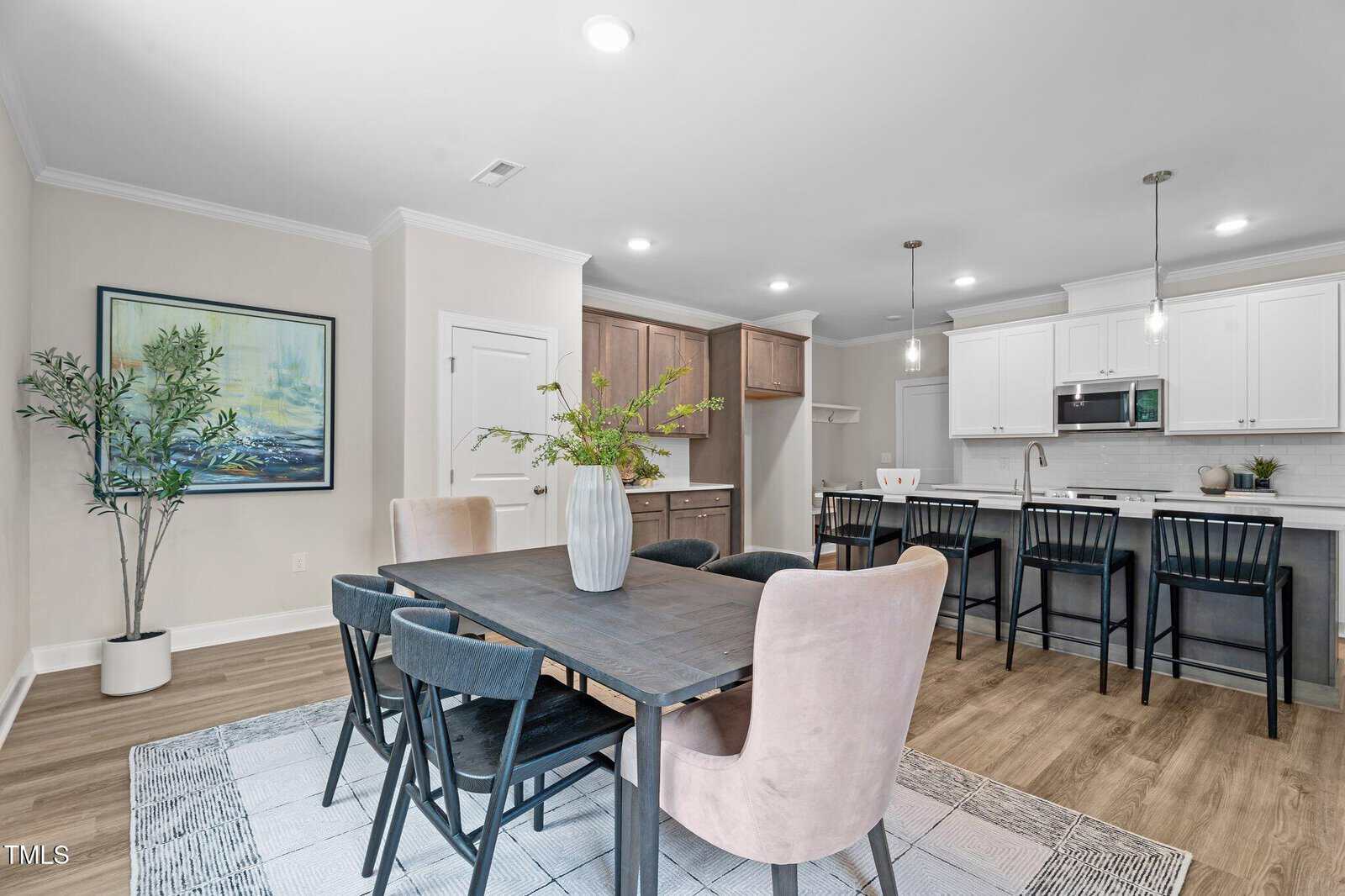Modern kitchen and dining area with a dark wood table, six chairs, pendant lights over a breakfast bar, potted plants, white cabinets, and light wood floors. A large framed artwork hangs on the wall.