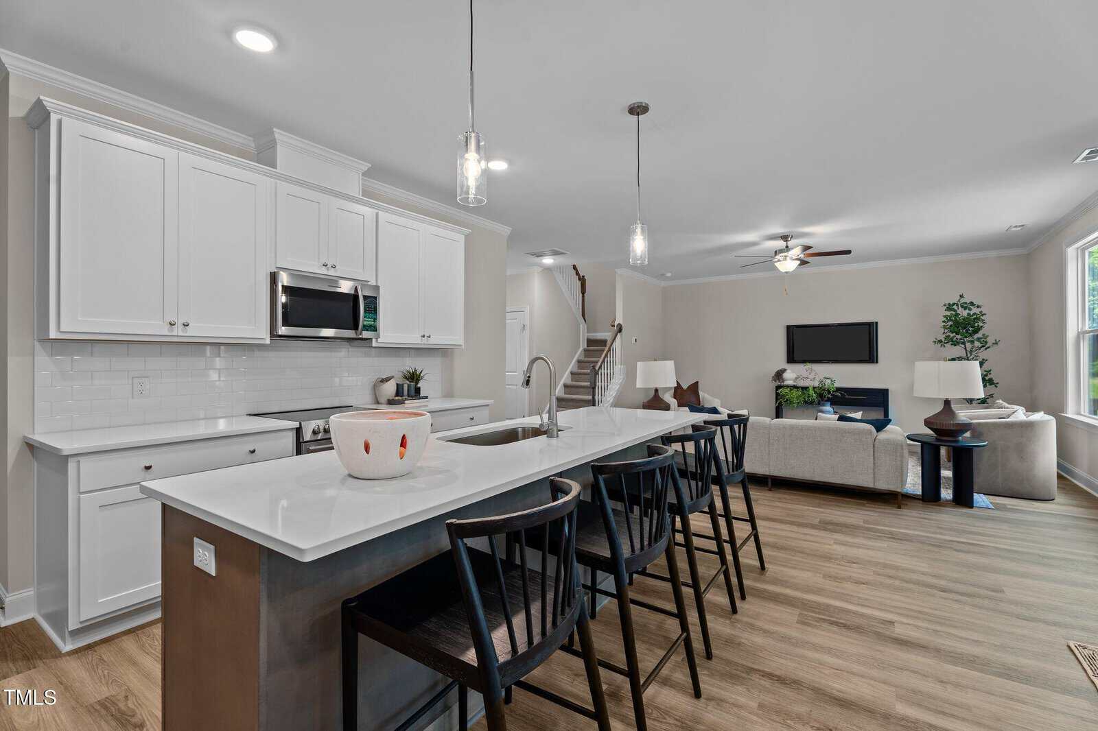 Modern kitchen with white cabinets, stainless steel appliances, and a large island with four black bar stools. The space opens to a living area with a couch, TV, and stairs leading upstairs. Natural light comes in through a window.