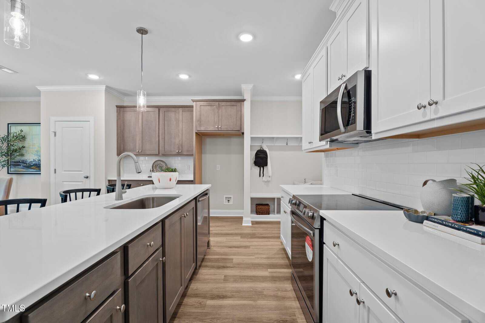 Modern kitchen with white cabinets, stainless steel appliances, a center island with a sink, light wood floors, pendant lighting, and a built-in nook with hooks and storage cubbies on the right wall.