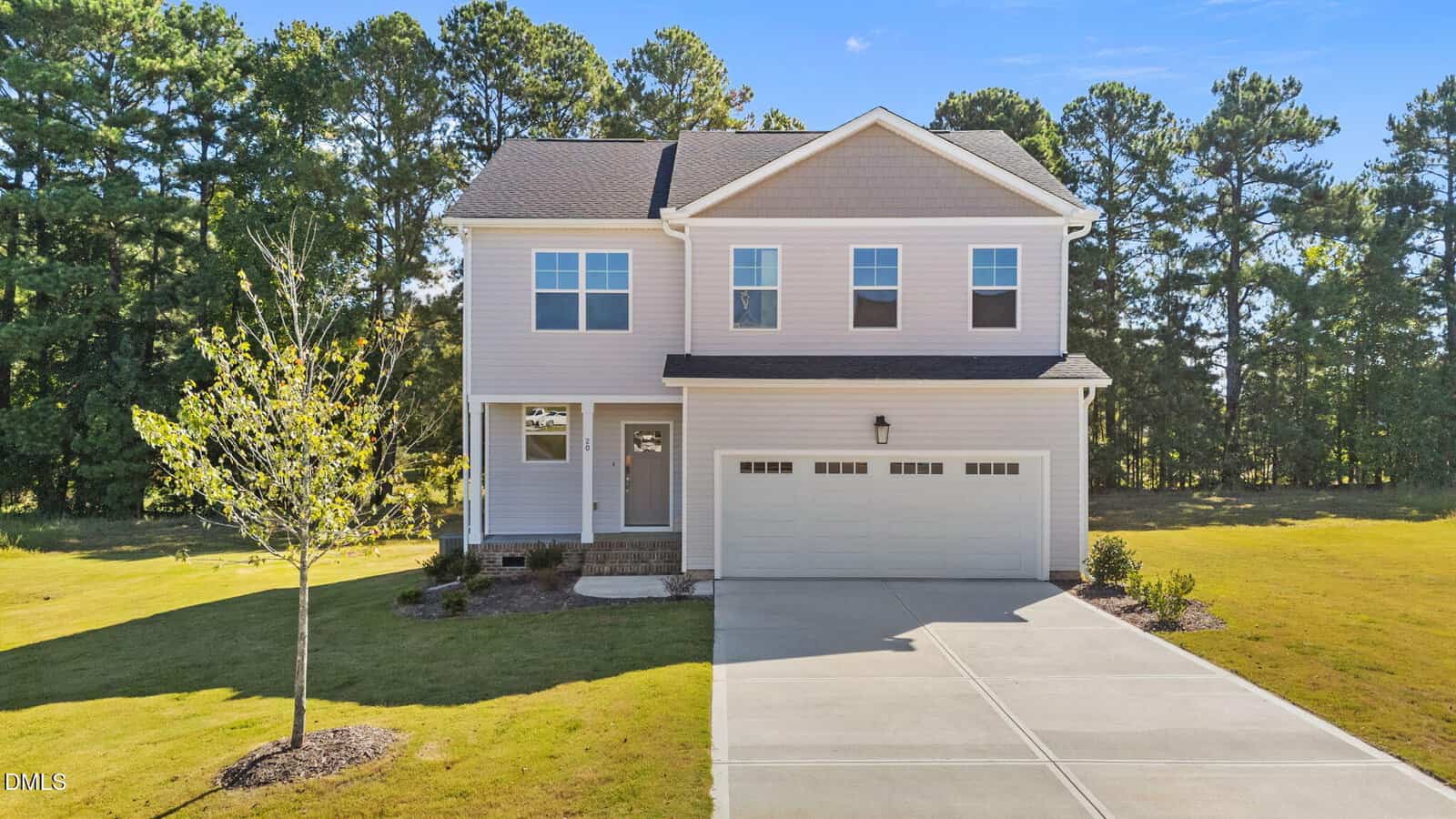 Charming two-story beige house at 20 Diamond Creek Drive, Zebulon, featuring a double garage, concrete driveway, and small front porch. A single young tree and minimal landscaping grace the front yard; the background is framed by a line of tall, green trees.