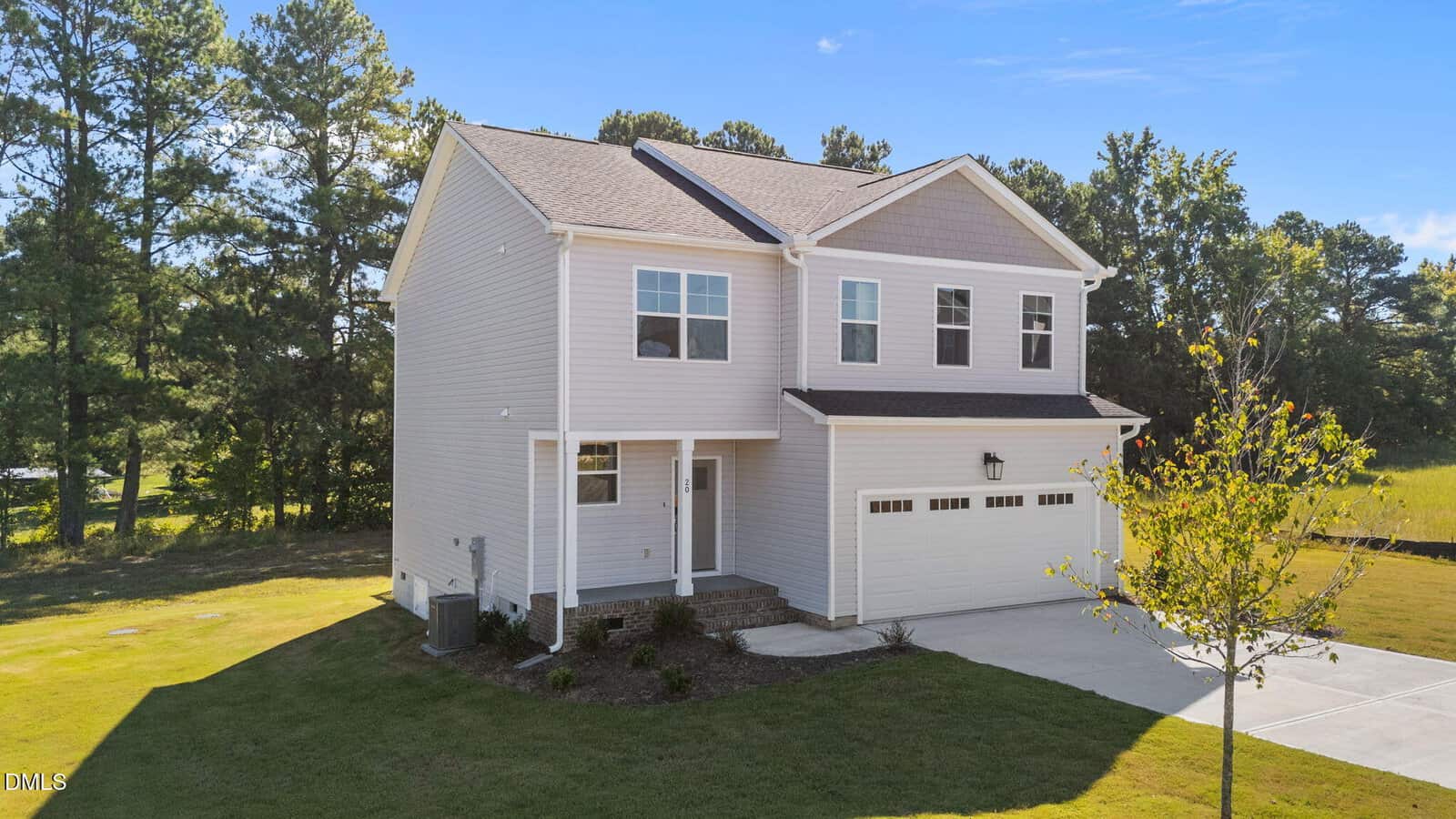 Two-story beige house with a double garage, small front porch, and neatly landscaped yard, set on a large green lot bordered by trees under a clear blue sky.